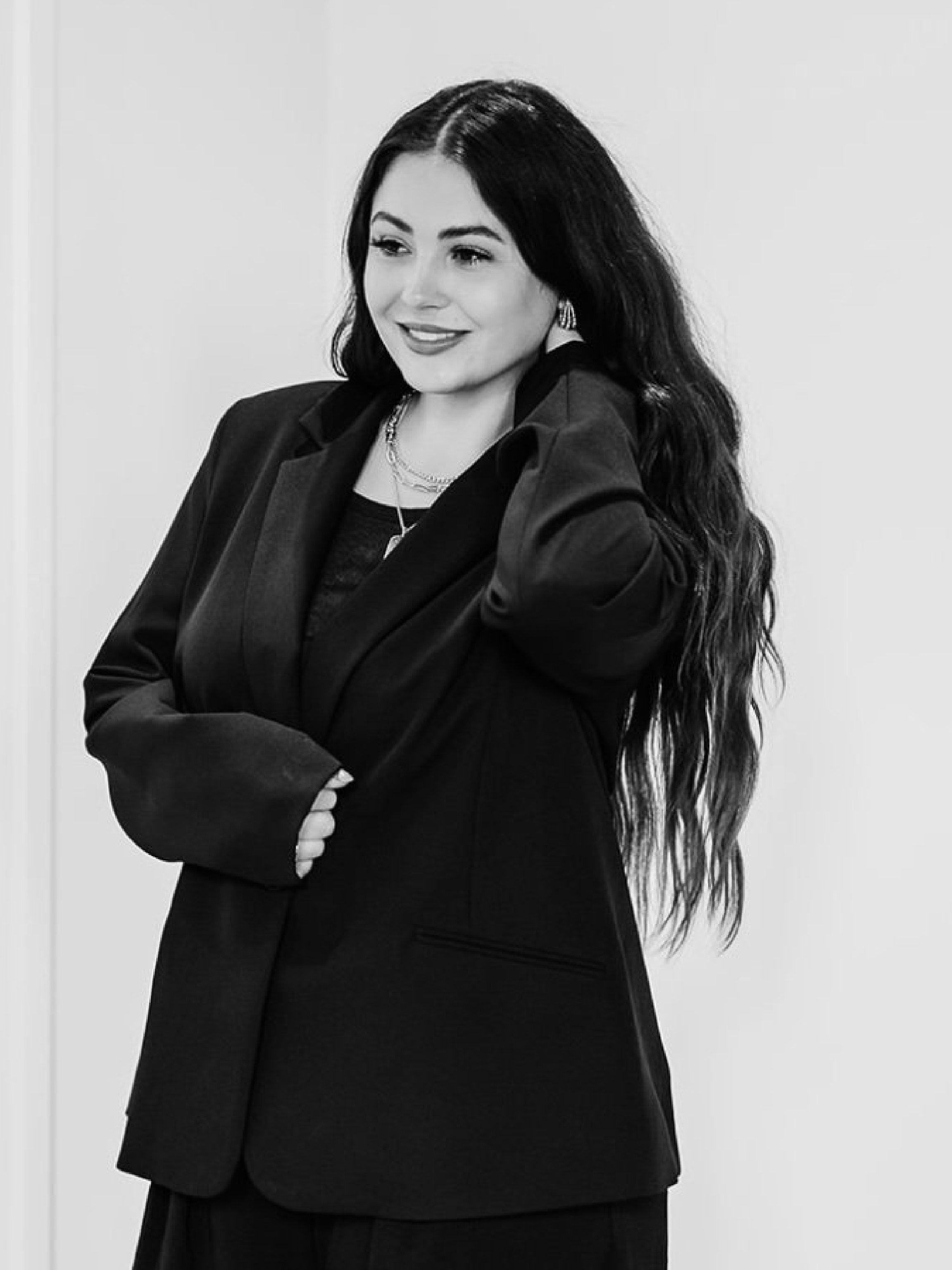 Black and white photo of a woman with long wavy hair, wearing a blazer, smiling, with an empty white wall background.