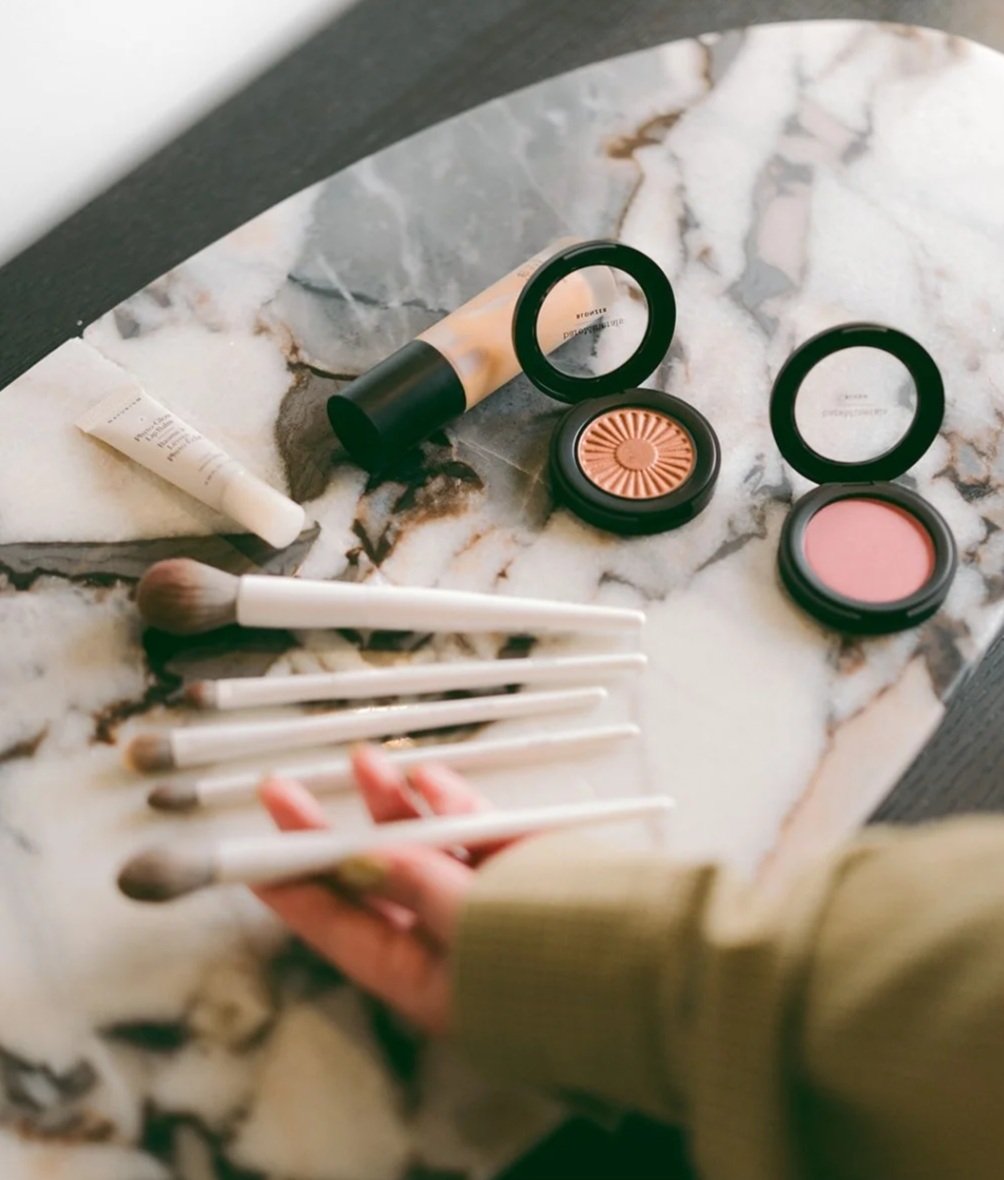 Beauty products on a marble surface, including foundation, blush, peach-colored eyeshadow, various makeup brushes, and a hand holding a few of the brushes.