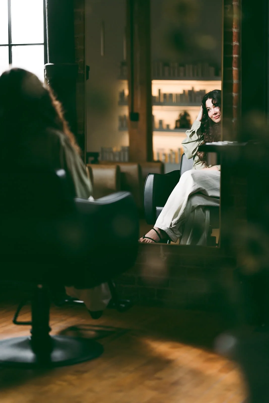 A woman with long curly hair sitting in a cafe, smiling, looking at someone off-camera. She is dressed in light-colored clothing and sandals, with a relaxed pose, next to bookshelves filled with books.