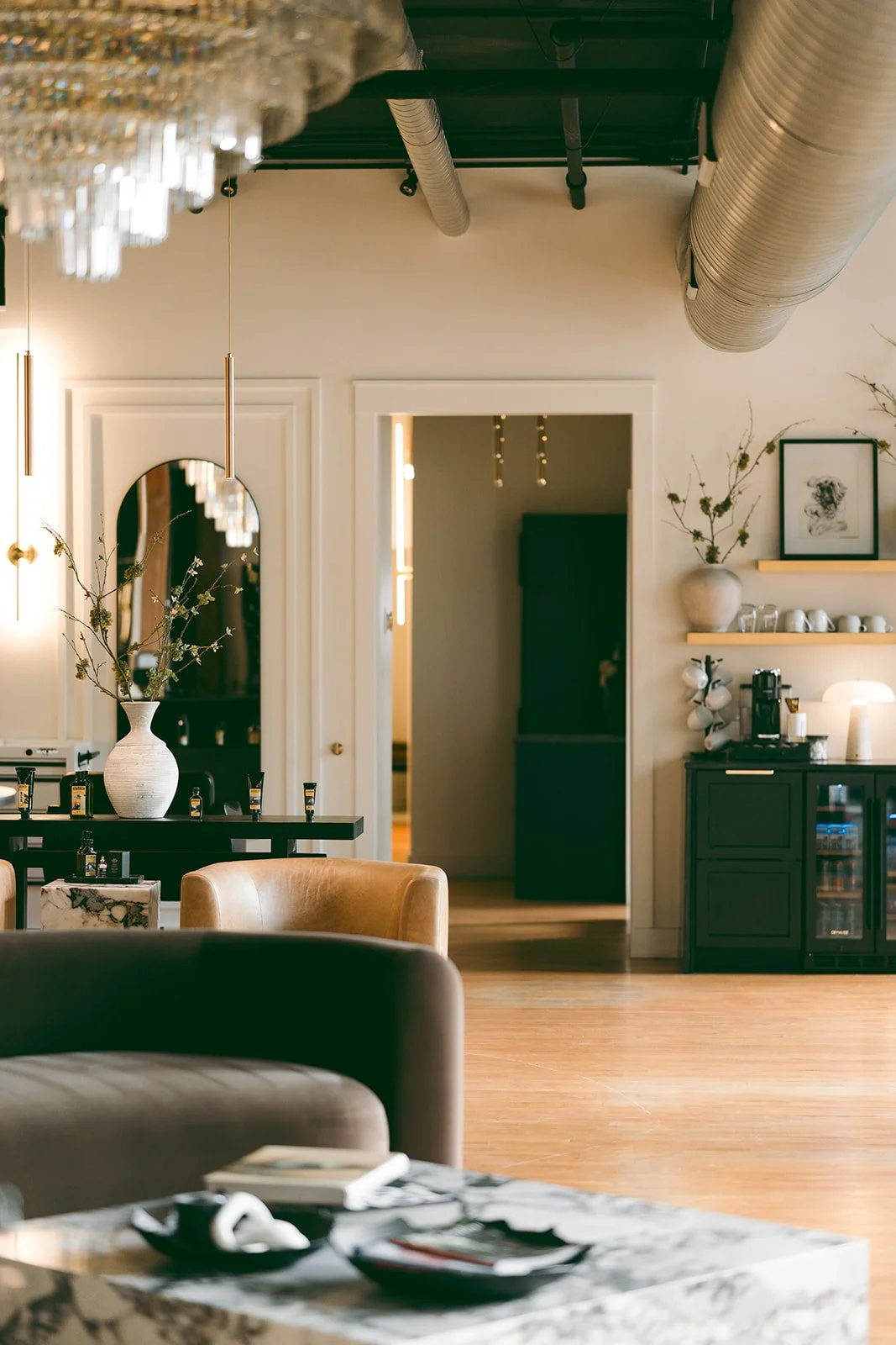 Interior of a modern cafe or lounge with a marble table, tan chairs, a black cabinet with a coffee machine, and decorative shelves with vases and framed artwork. Ductwork is visible on the ceiling.