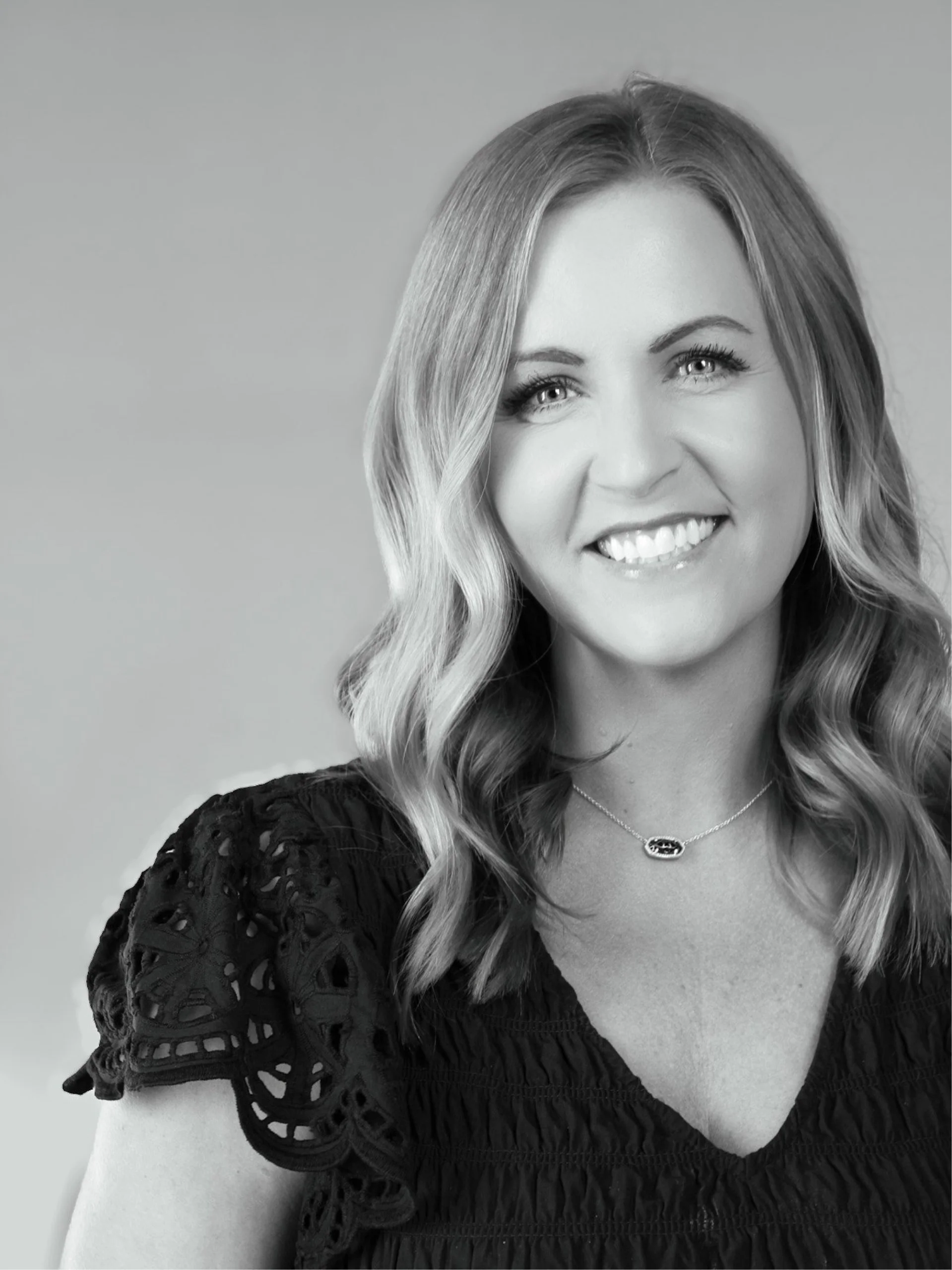 A woman with long wavy hair smiling, wearing a black lace top and a necklace with a small pendant, seen from the shoulders up.