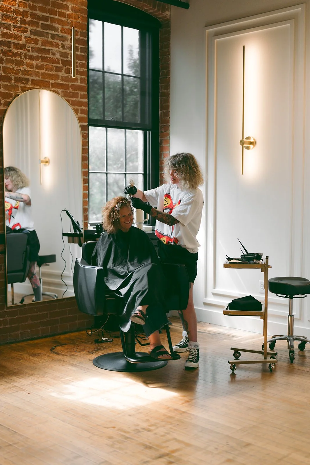 A hairstylist with curly hair and tattoos on her arms is applying hair dye or highlights to a client's curly hair in a salon with a large mirror, brick wall, and window. The client is seated in a salon chair, smiling, wearing a black cape. The salon has wooden floors, decorative wall lighting, and a mobile table with hair tools.