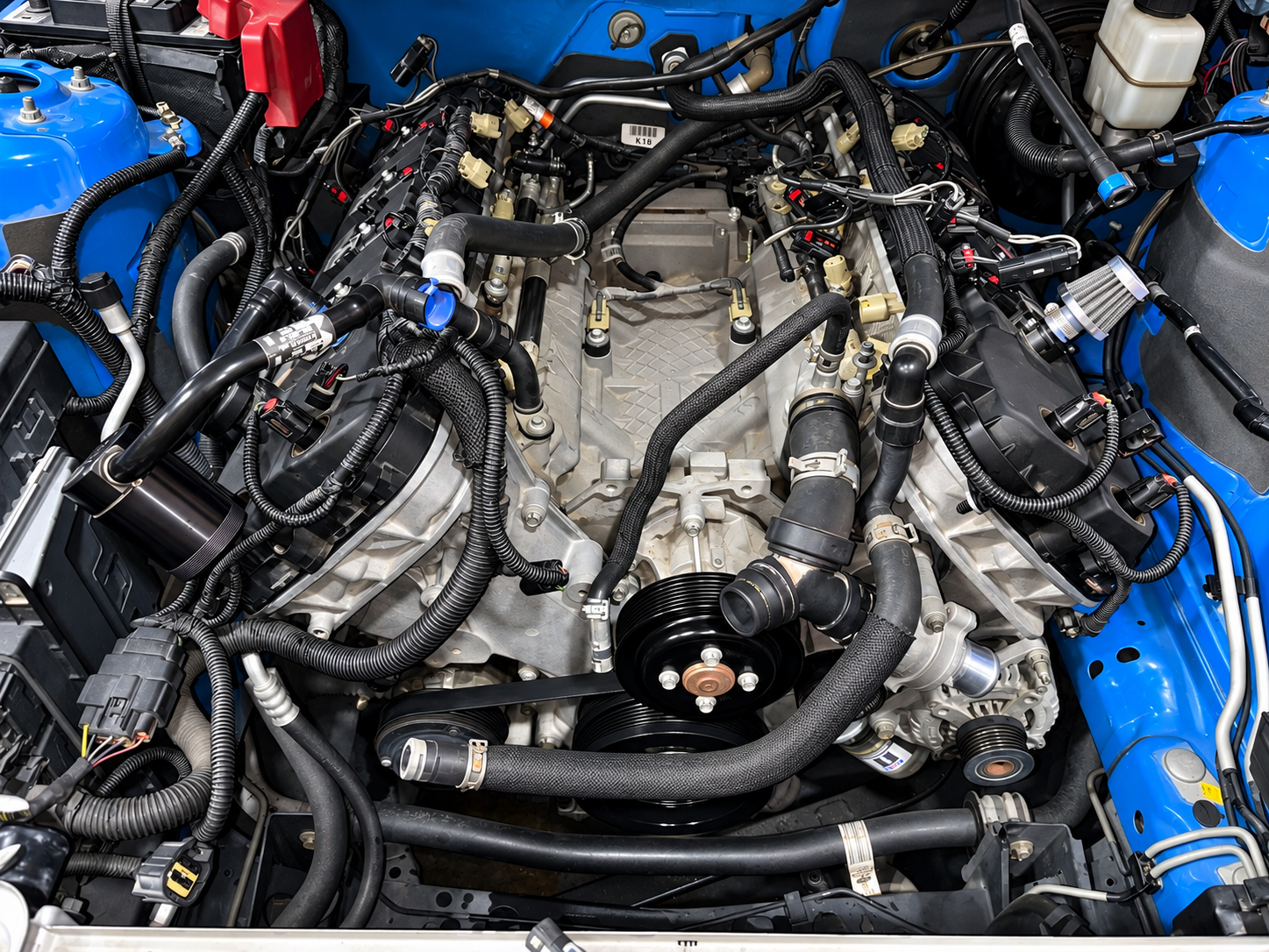 Close-up view of a car engine with various black hoses, belts, and metallic components, with a blue car body surrounding the engine bay.