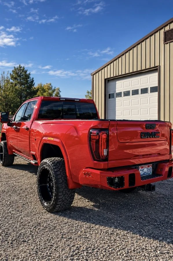 Red GMC pickup truck parked outdoors on gravel near a metal building with a white garage door under a blue sky with scattered clouds.