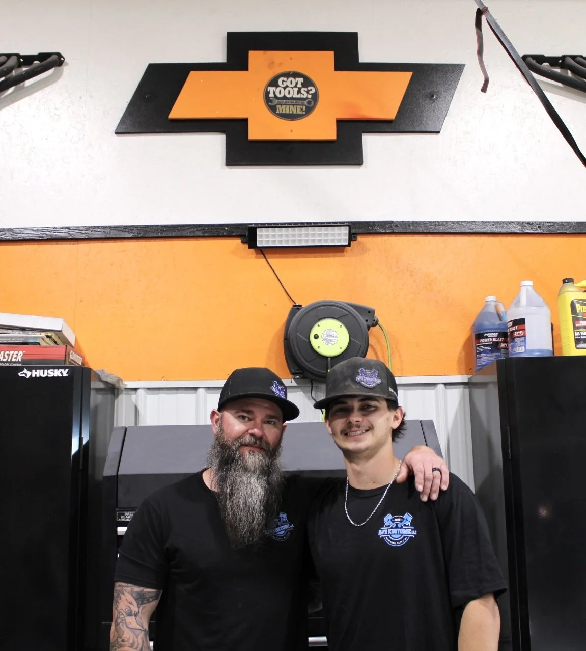 Two men in black shirts and black caps smiling, standing in a garage with a Chevrolet logo sign above them and a wall with tools and supplies, including motor oil bottles, behind them.
