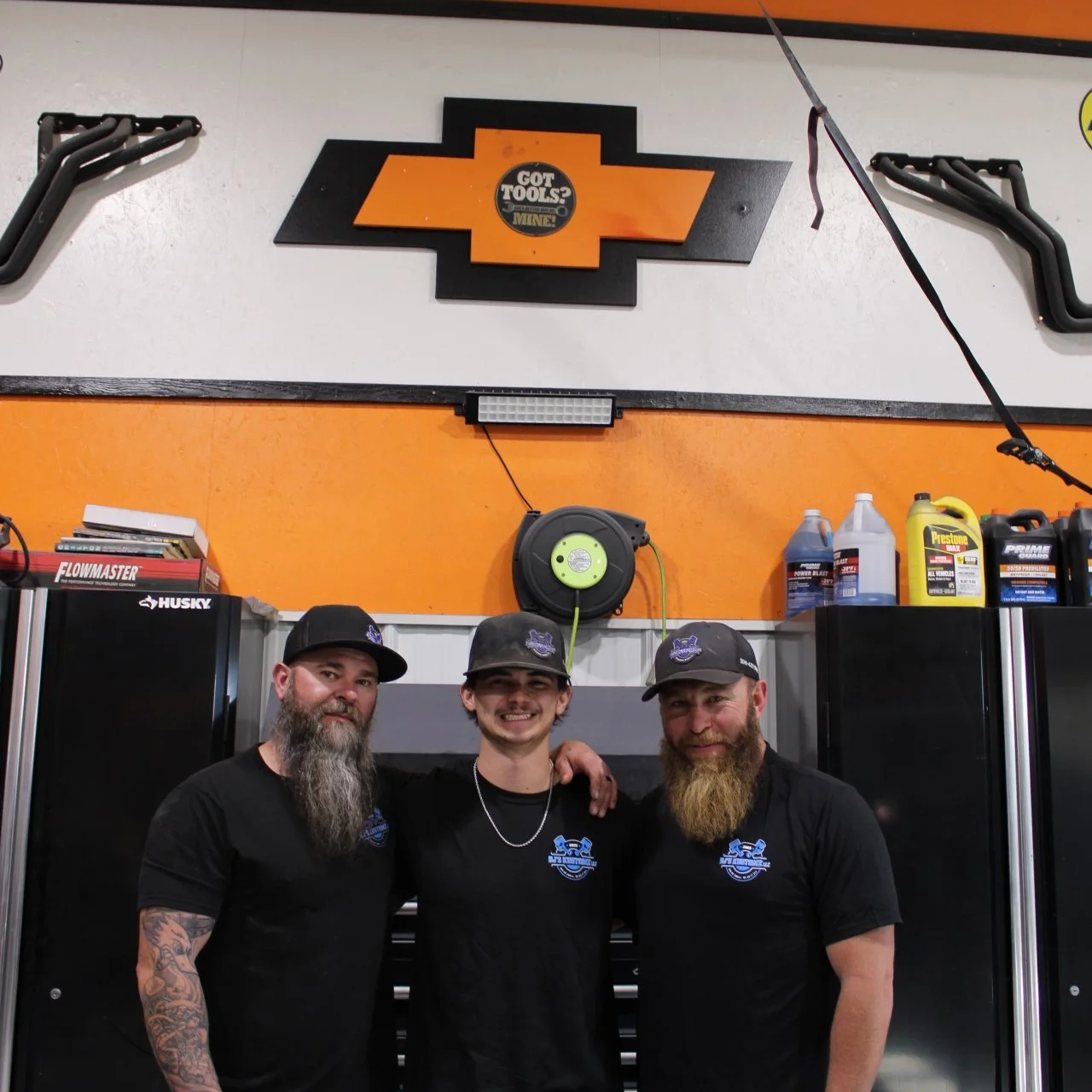 Three men wearing black shirts and caps, standing in a garage. Two of them have beards and the man on the left has colorful tattoos on his arm. They are smiling and have their arms around each other. There are automotive tools and products in the background, with a Chevrolet logo sign and orange and black decor.