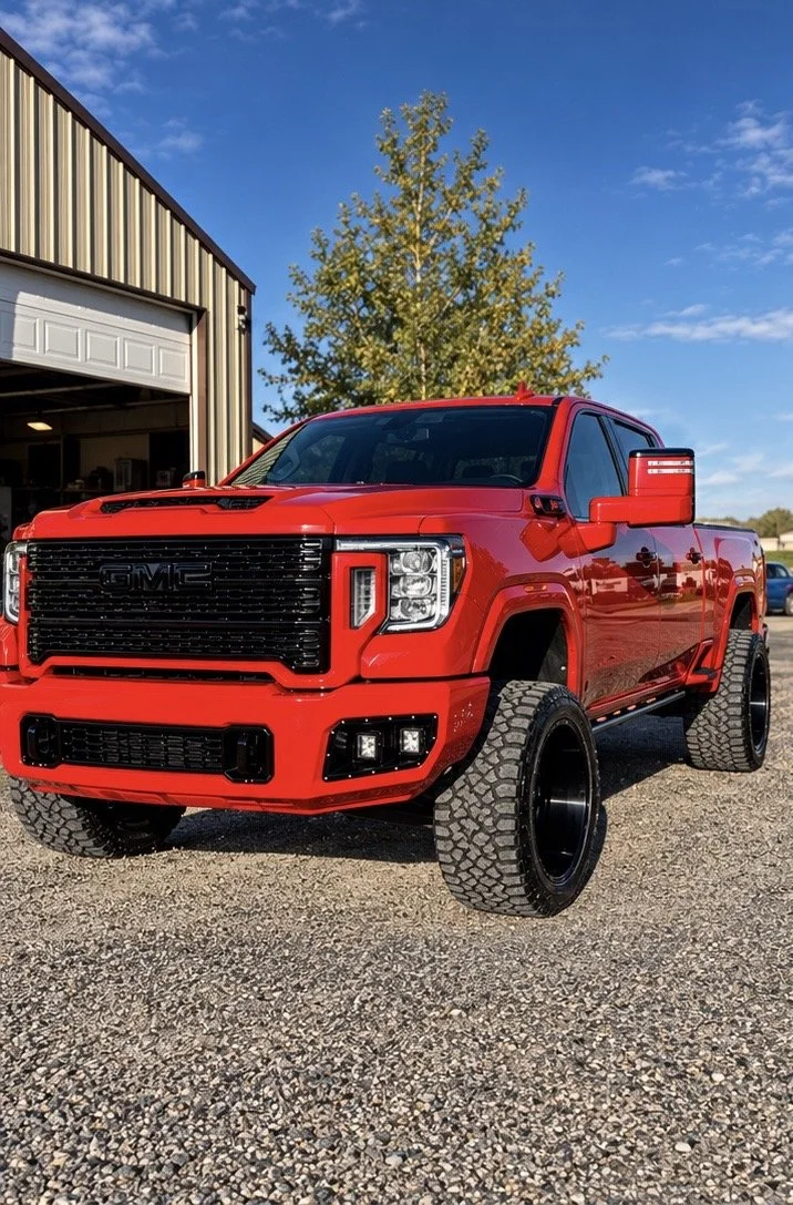Red GMC pickup truck with large tires parked outside near a barn and a tree under a blue sky.