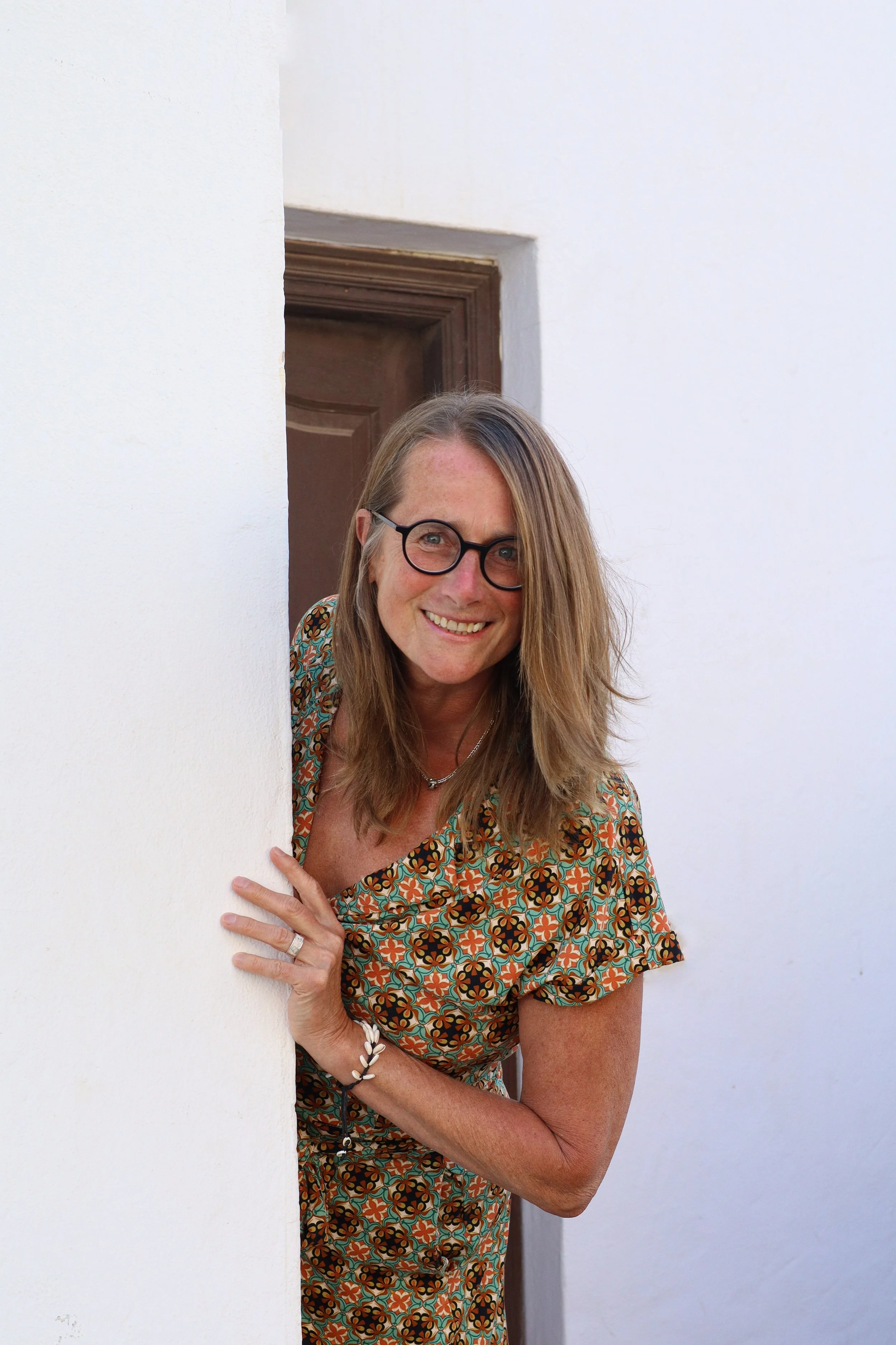Barbara mit Brille, blonden, langen Haaren, trägt ein bunt gemustertes Kleid, schaut neugierig durch eine Türöffnung, steht vor weißer Wand.
