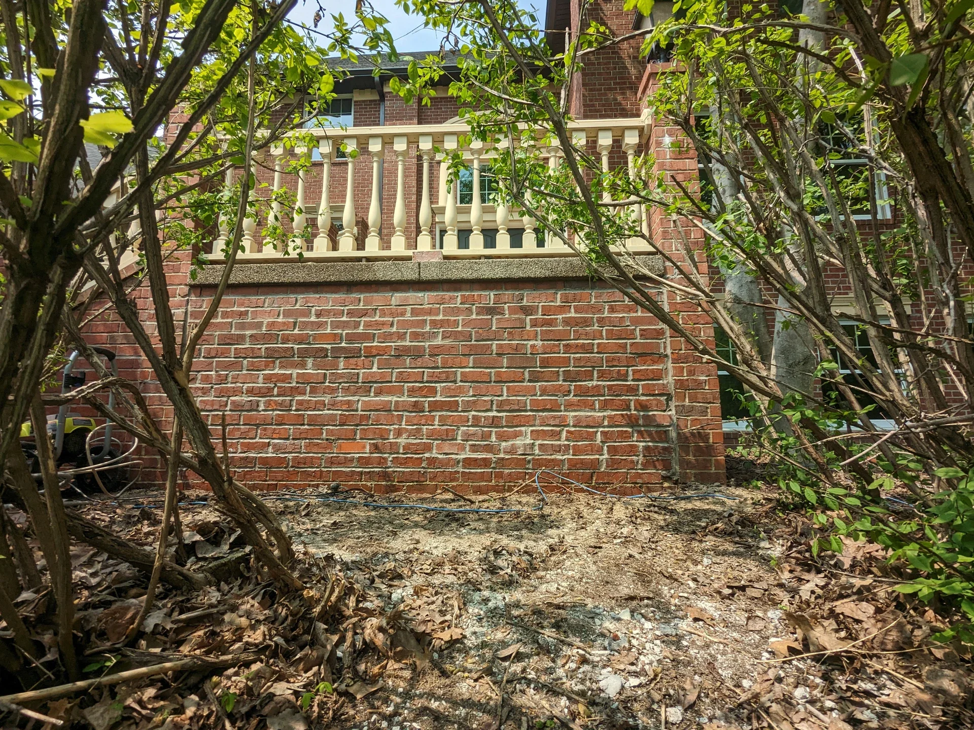 View of a brick house with a stone balcony and white railing, seen from a ground-level perspective through bushes and trees with leaning branches and dried leaves.