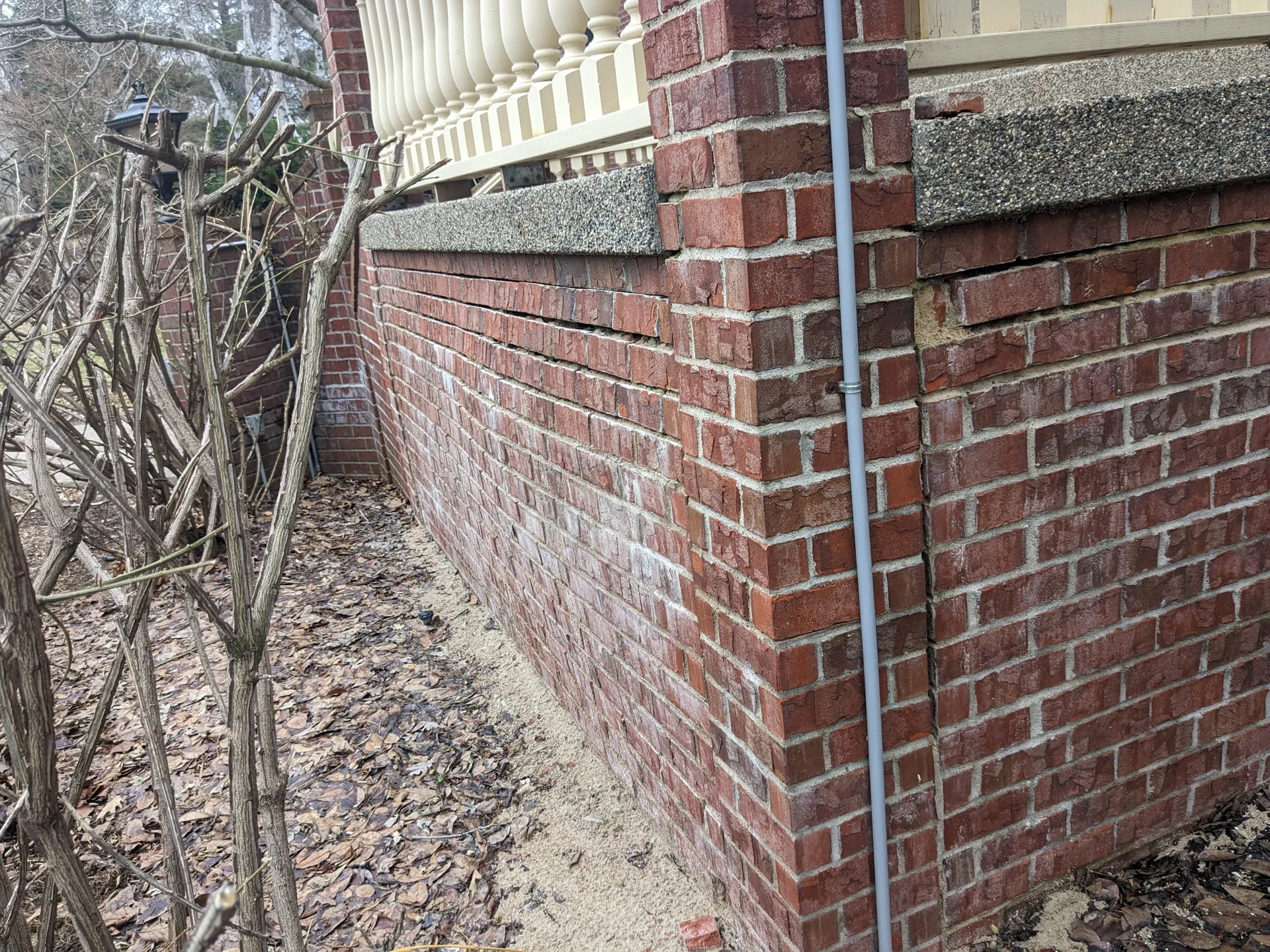 Corner of a brick house with a downspout, surrounded by fallen leaves and bare shrub branches.