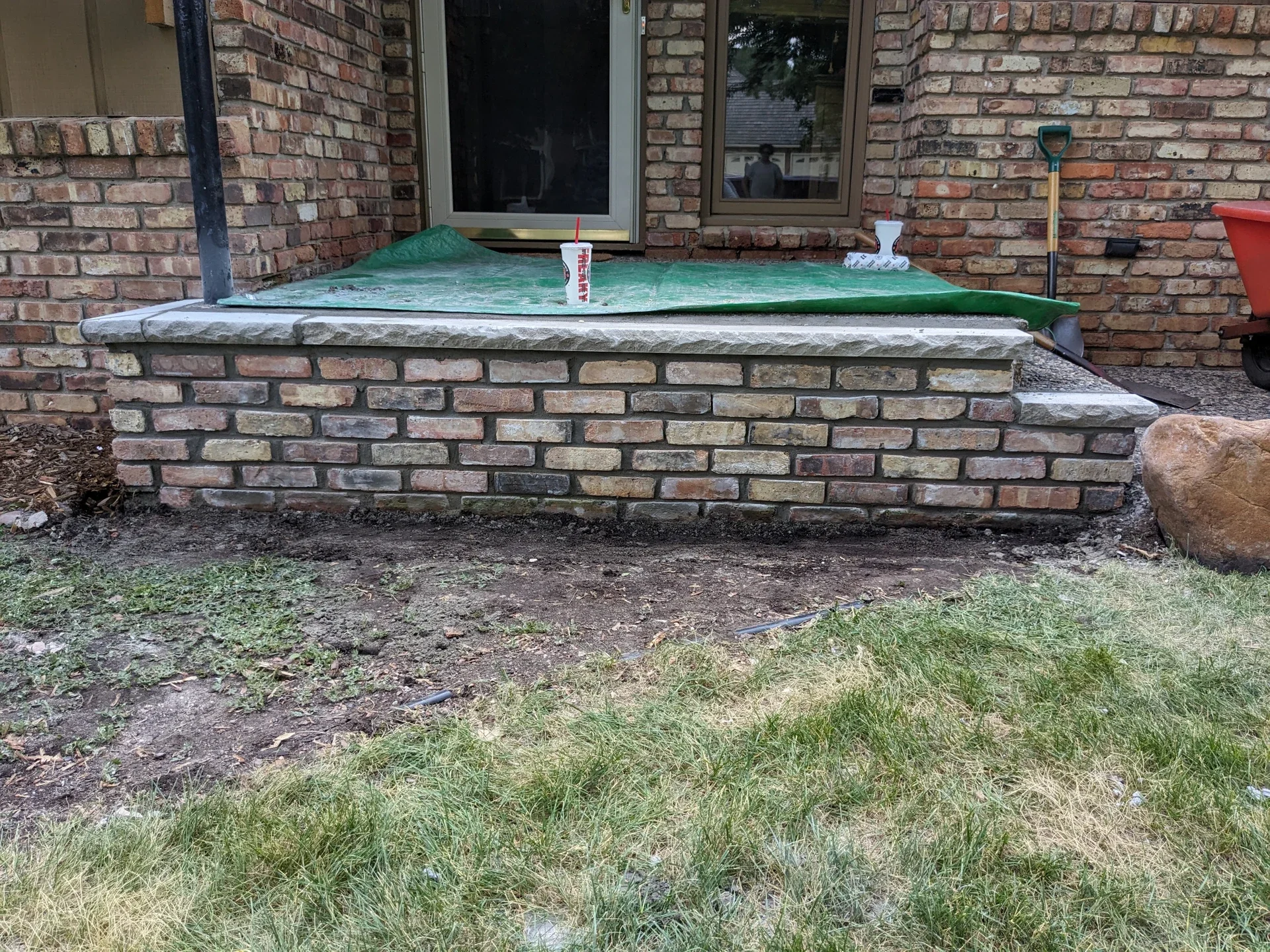 A brick porch with a green tarp cover, a cup with a straw, a shovel, and a wheelbarrow near a brick house.