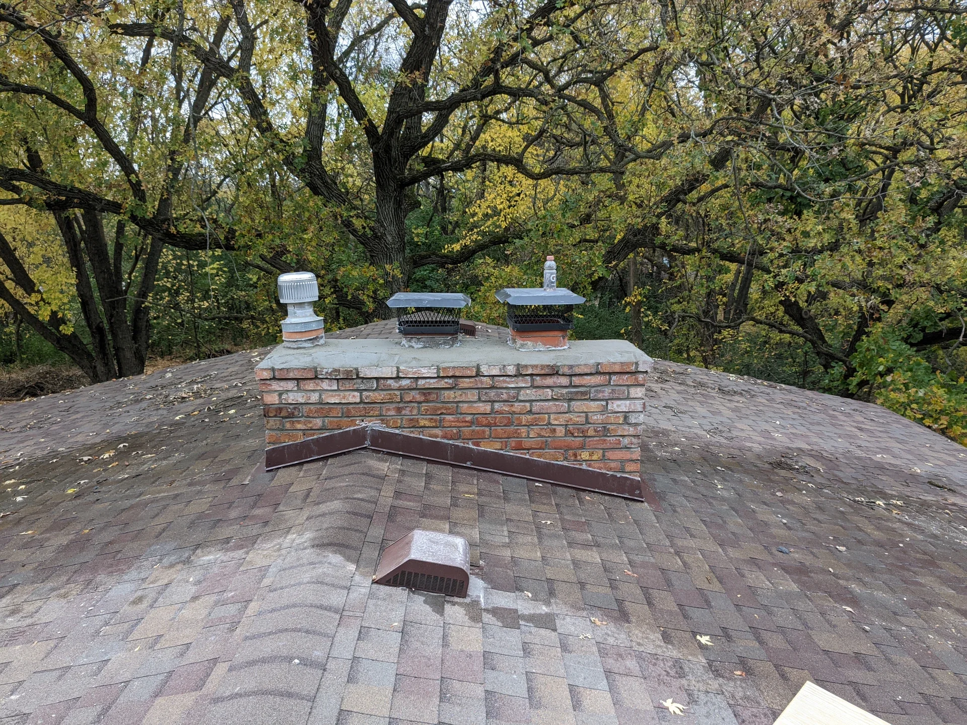 View of a house roof with a brick chimney, ventilation and exhaust vents, surrounded by trees with autumn leaves.