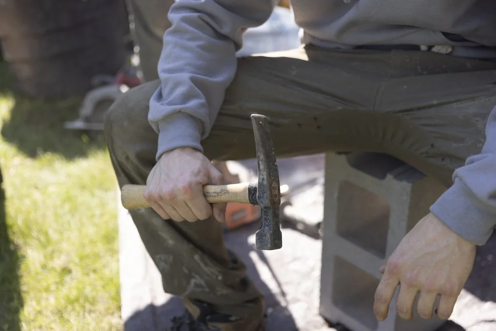 A person sitting outdoors on a cinder block, holding a hammer in their right hand, with a grassy area and trees in the background.
