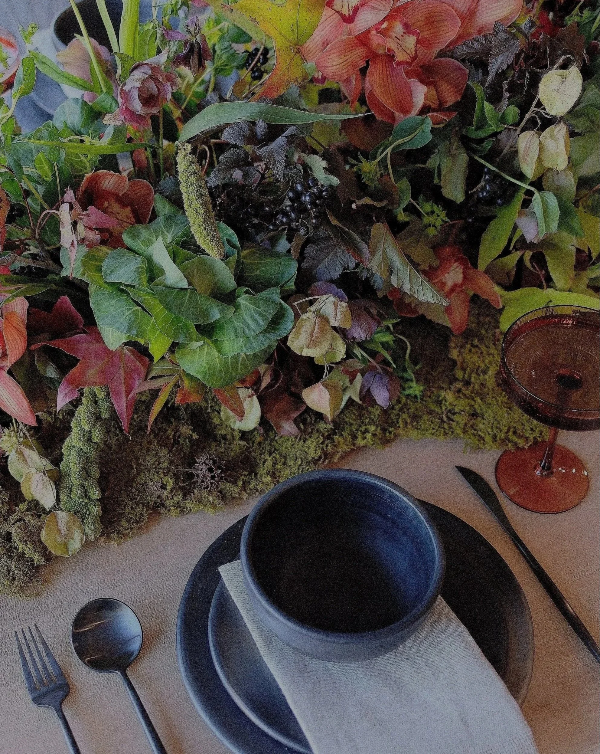 Elegant table setting with black plates, silverware, and a centerpiece of colorful flowers, leafy greens, berries, and moss