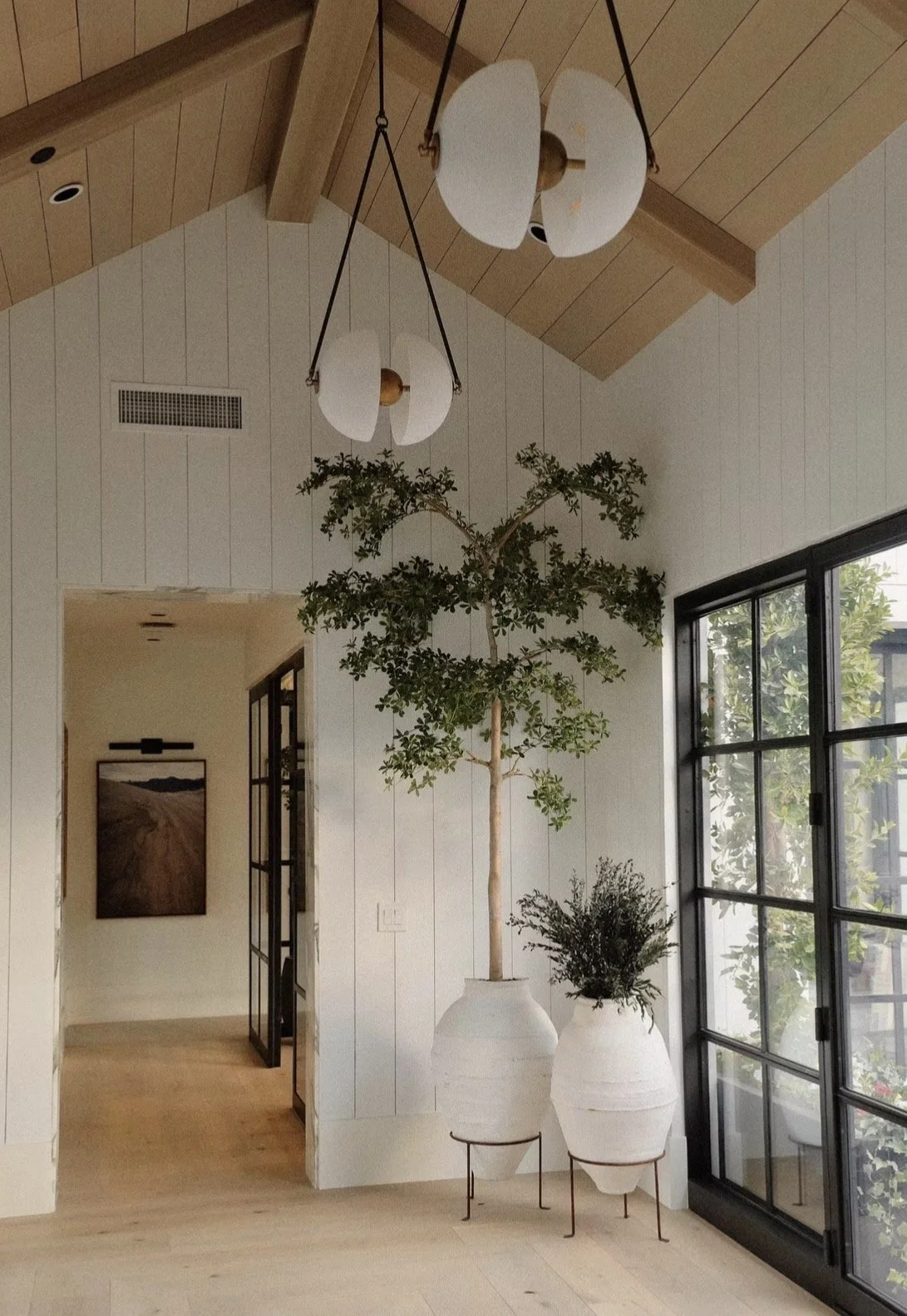 Modern home interior with white-paneled walls, exposed wood-beam ceiling, pendant lights, large potted trees, and black-framed windows.