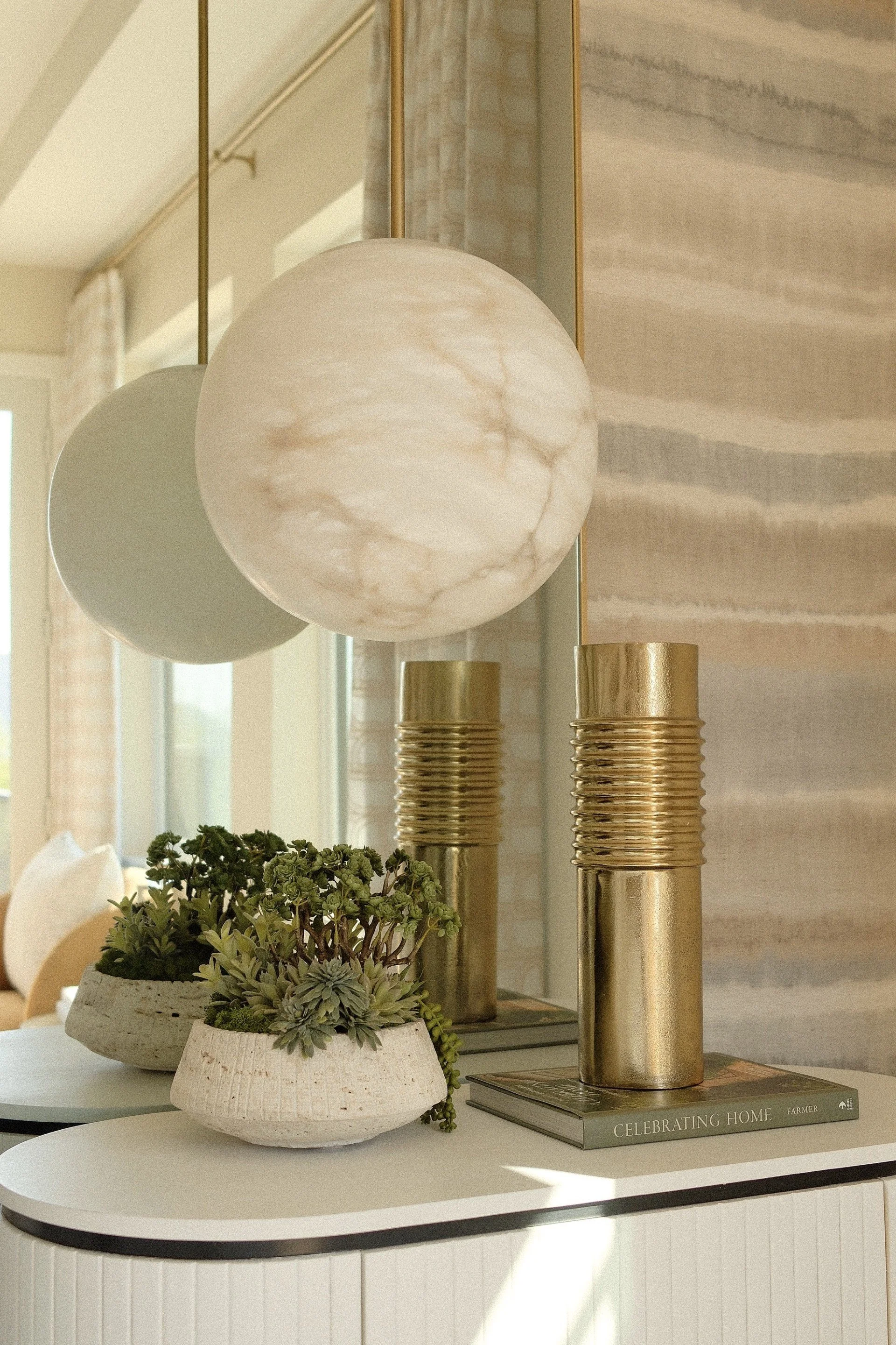 Modern console table styled with brass lamps, a white globe pendant light, potted succulents, and stacked books.