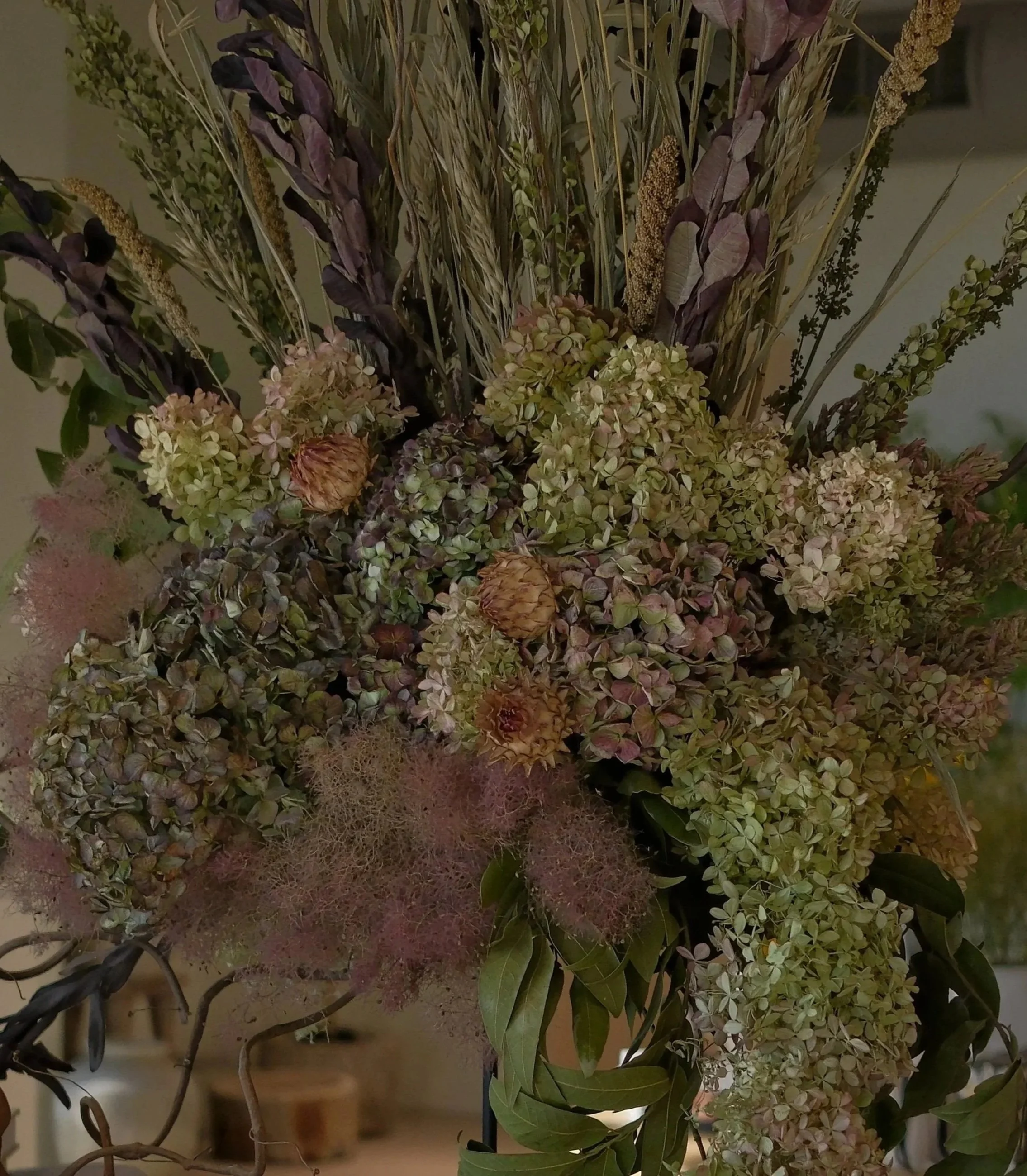 Large dried floral arrangement with muted green and purple hydrangeas, tall grasses, seed pods, and leafy stems