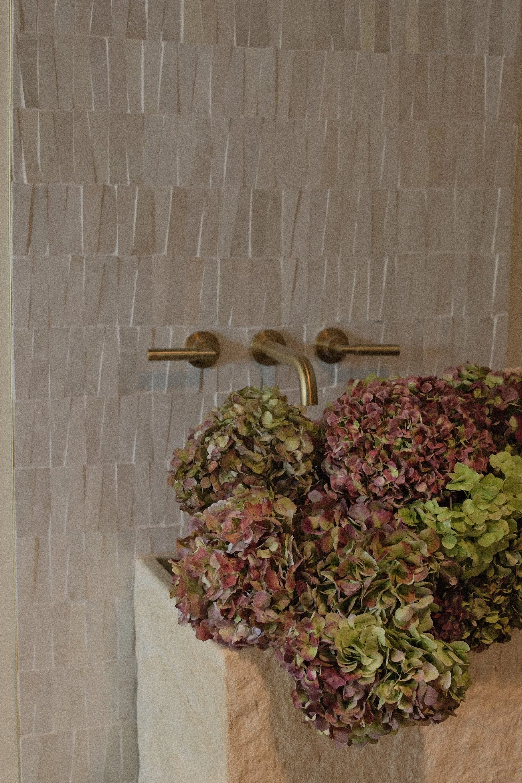 Modern bathroom with white textured tile, brushed brass wall-mounted faucet, and colorful dried hydrangeas in a vessel