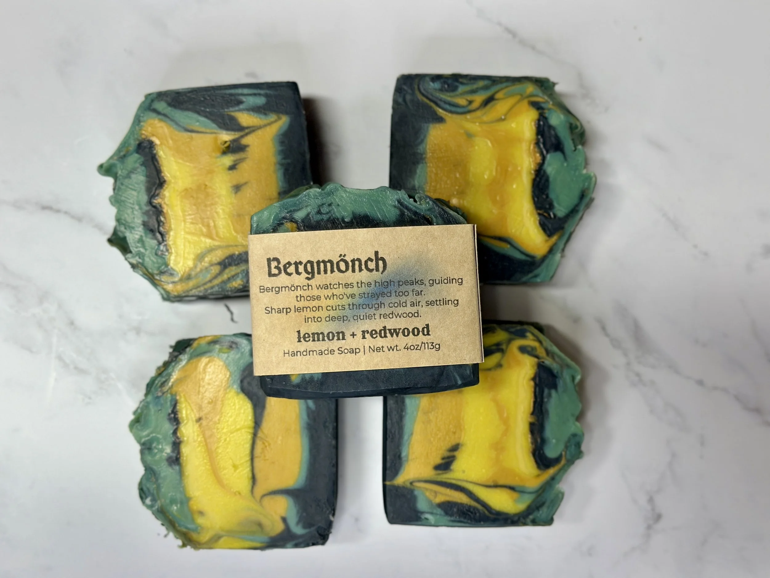 Photo of handcrafted artisan soaps, posed to appeal. One soap shows a label with the soap's name, Bergmönch. The soaps include bright and muted yellows, teal, and deep blue.