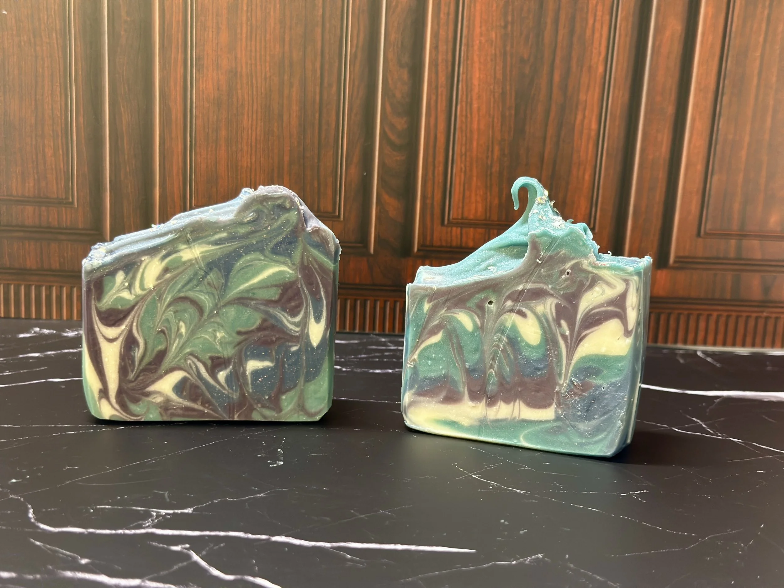 Photo of handcrafted artisan soaps, posed to appeal. The soaps are made of deep blue, teal, purple, and lighter blue swirls, topped with biodegradable glitter.