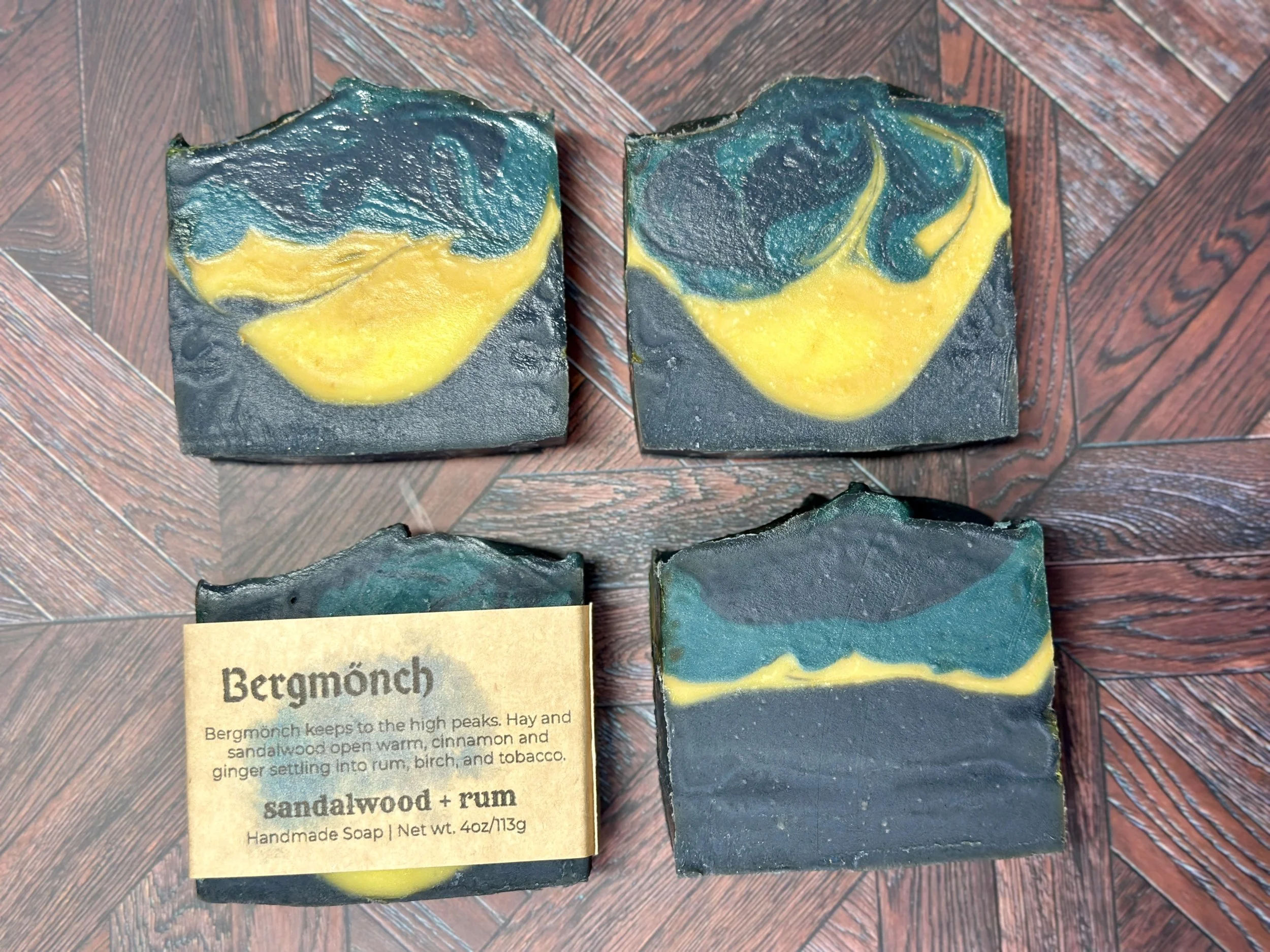 Photo of handcrafted artisan soaps, posed in a four-quadrant layout. One soap shows a label with the soap's name, Bergmönch. The soaps include bright and muted yellows, teal, and deep blue.