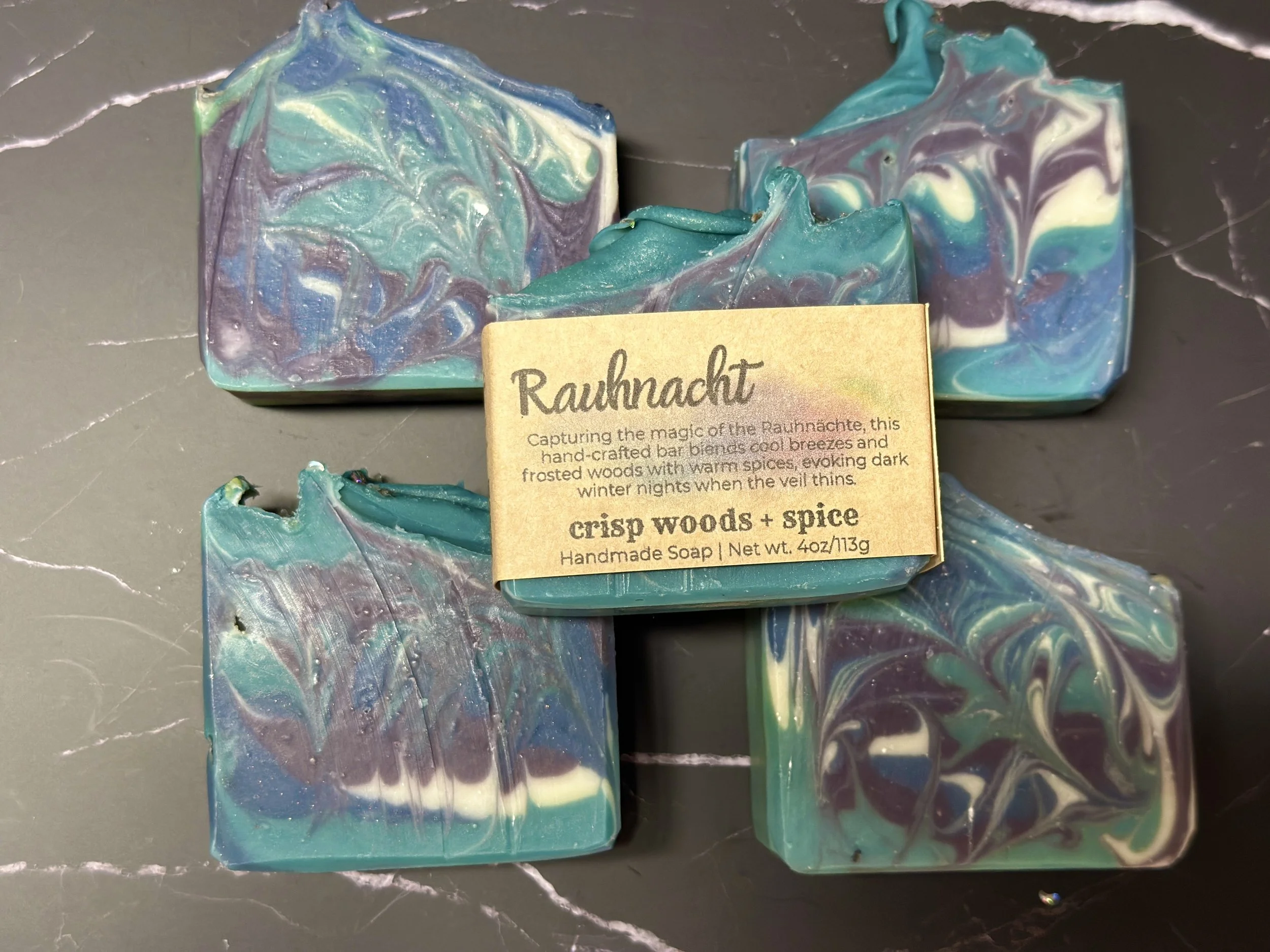 Photo of handcrafted artisan soaps, posed to appeal. The soaps are made of deep blue, teal, purple, and lighter blue swirls, topped with biodegradable glitter.