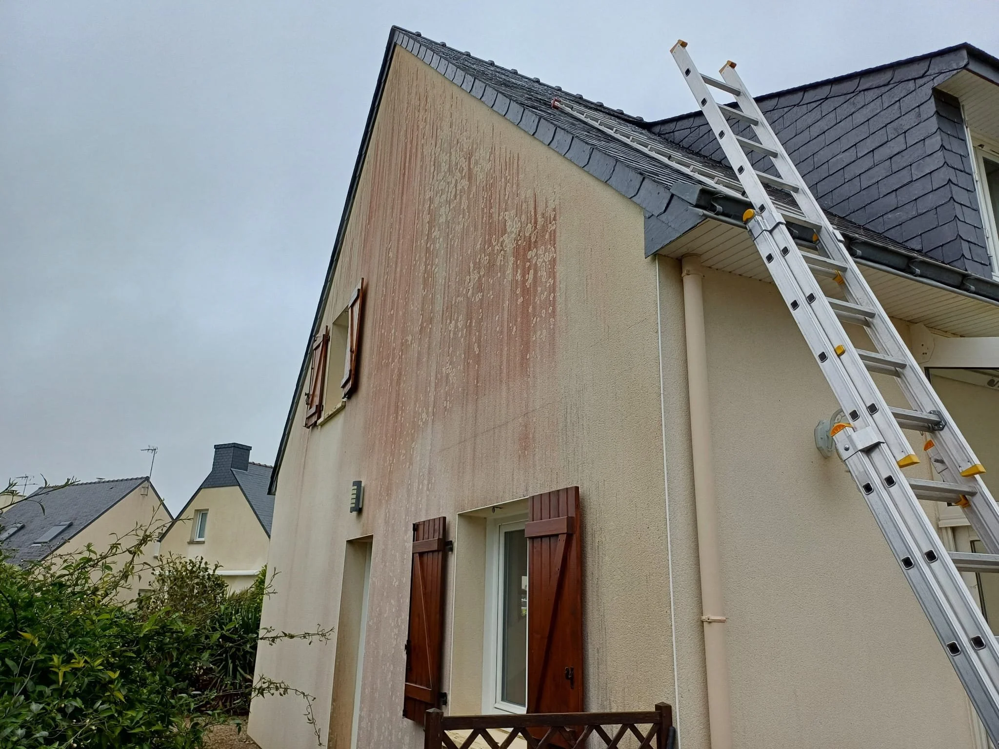 Une maison à deux étages avec un mur extérieur en cours de rénovation, une échelle en aluminium appuyée contre le toit et des volets en bois rouge au dessous des fenêtres. Il y a des végétaux verts au premier plan et un ciel nuageux.