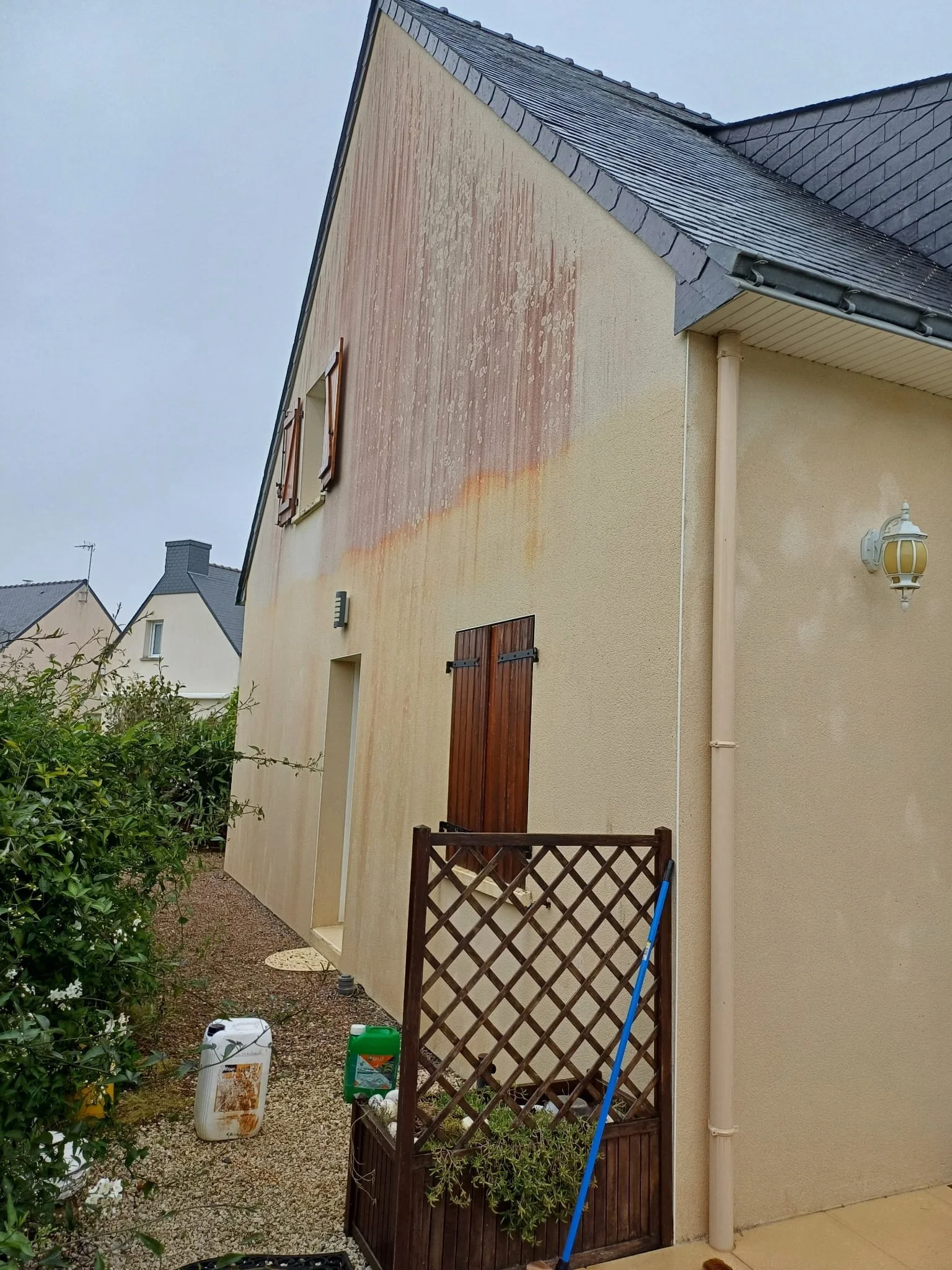Mur extérieur d'une maison en cours de nettoyage, avec des outils de nettoyage au sol et un mur partiellement lavé, sous un ciel gris.
