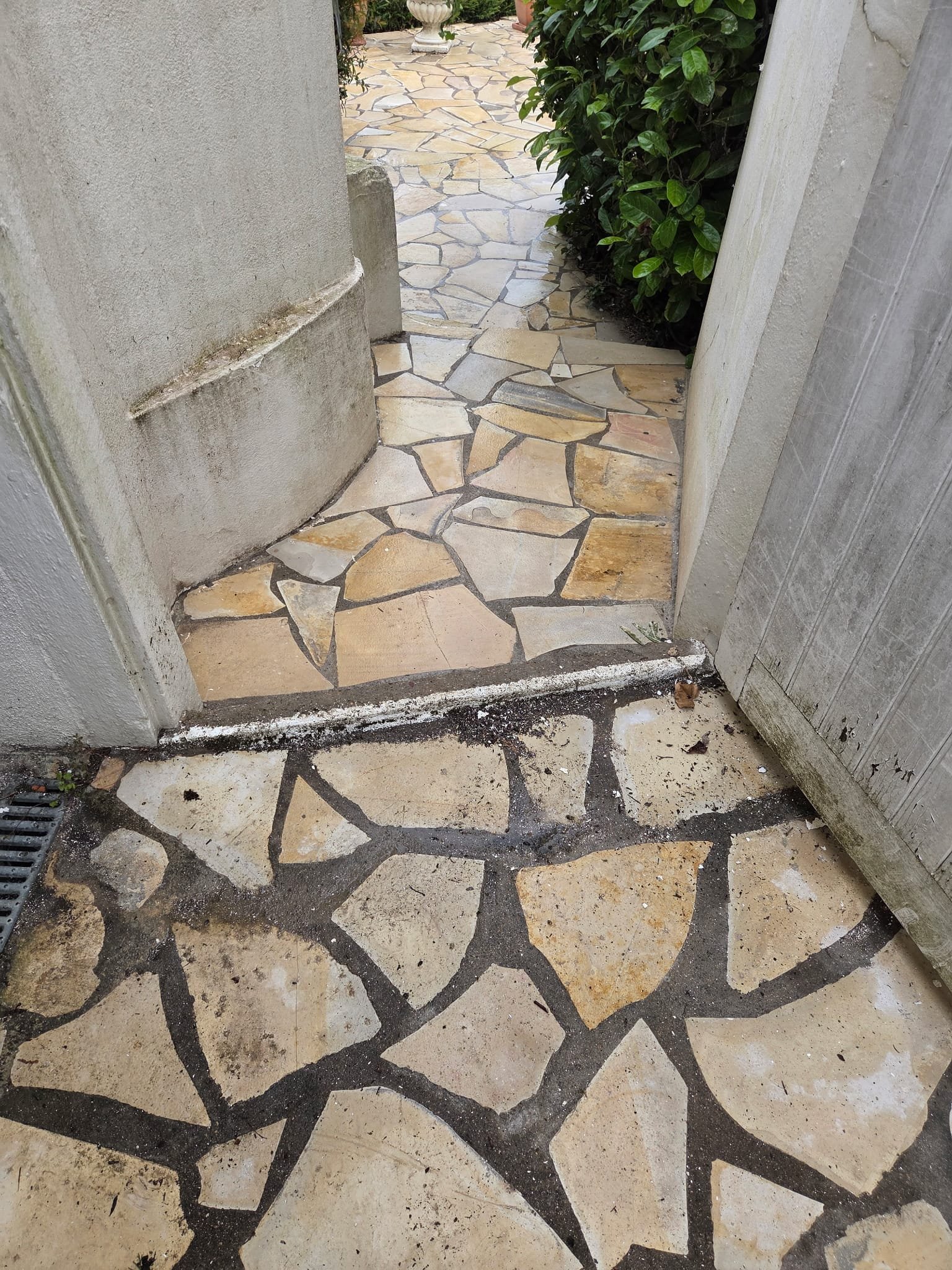 Une entrée avec un chemin en mosaïque de pierre entouré de murs en plâtre gris. Le chemin est composé de dalles de pierre beige clair avec des joints en mortier noir.