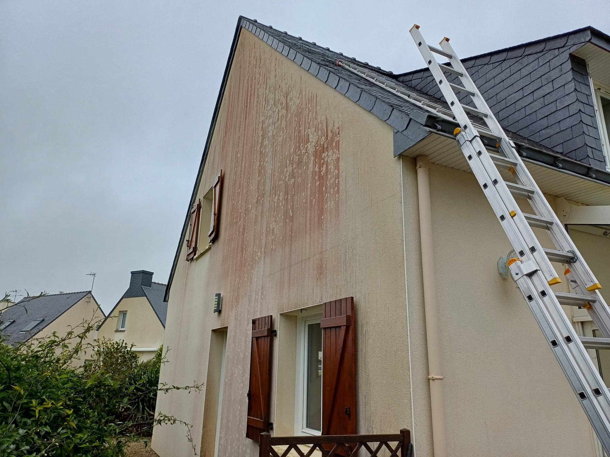 Une maison avec un mur en cours de rénovation, une échelle en position, des fenêtres avec volets en bois, un toit en tuiles et un ciel nuageux.
