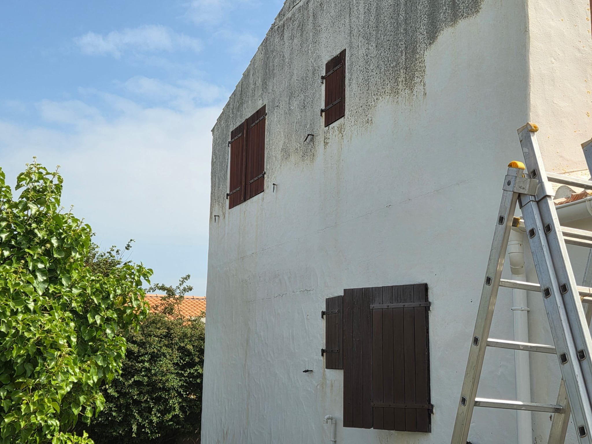 Une maison blanche avec deux fenêtres à volets en bois et une porte également en bois, une échelle en aluminium posée contre le mur, des arbres verts à gauche et un ciel bleu avec quelques nuages.