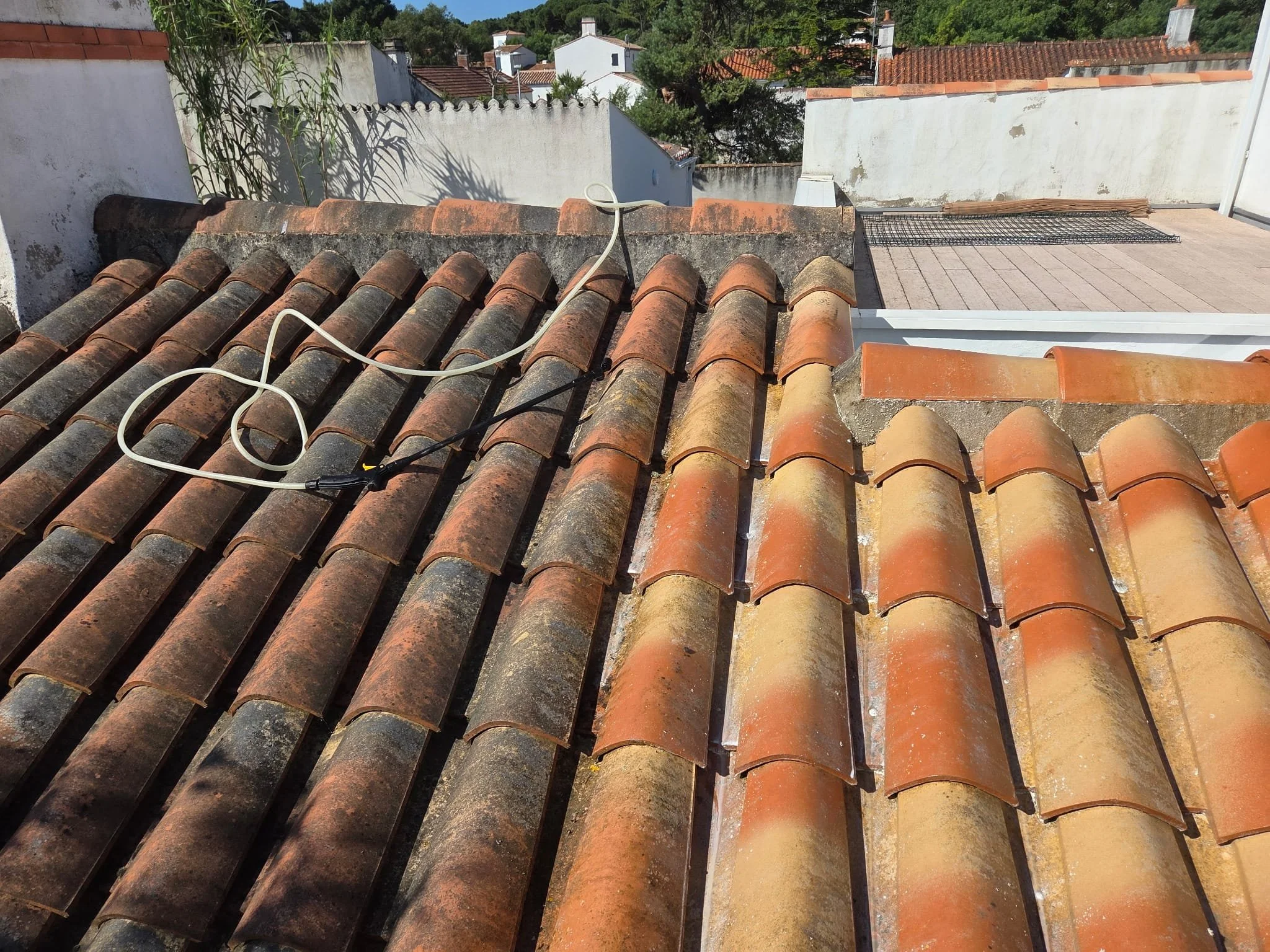 Vue d’un toit en tuiles en terre cuite, avec des câbles électriques visibles, des murs blancs environnants, un espace pour sécher ou stocker des objets, et des bâtiments avec des toits en tuiles rouges en arrière-plan sous un ciel ensoleillé.