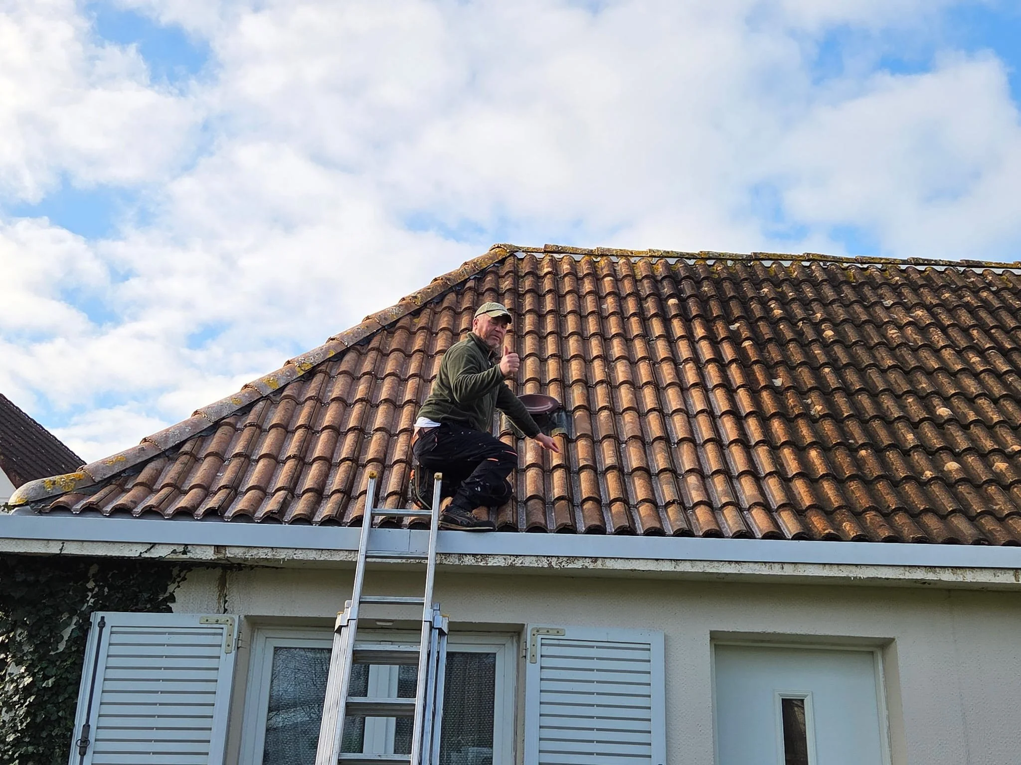 Un homme portant une veste verte et une casquette, debout sur une échelle, en train de travailler sur la toiture en tuiles d'une maison. Le ciel est partiellement nuageux.
