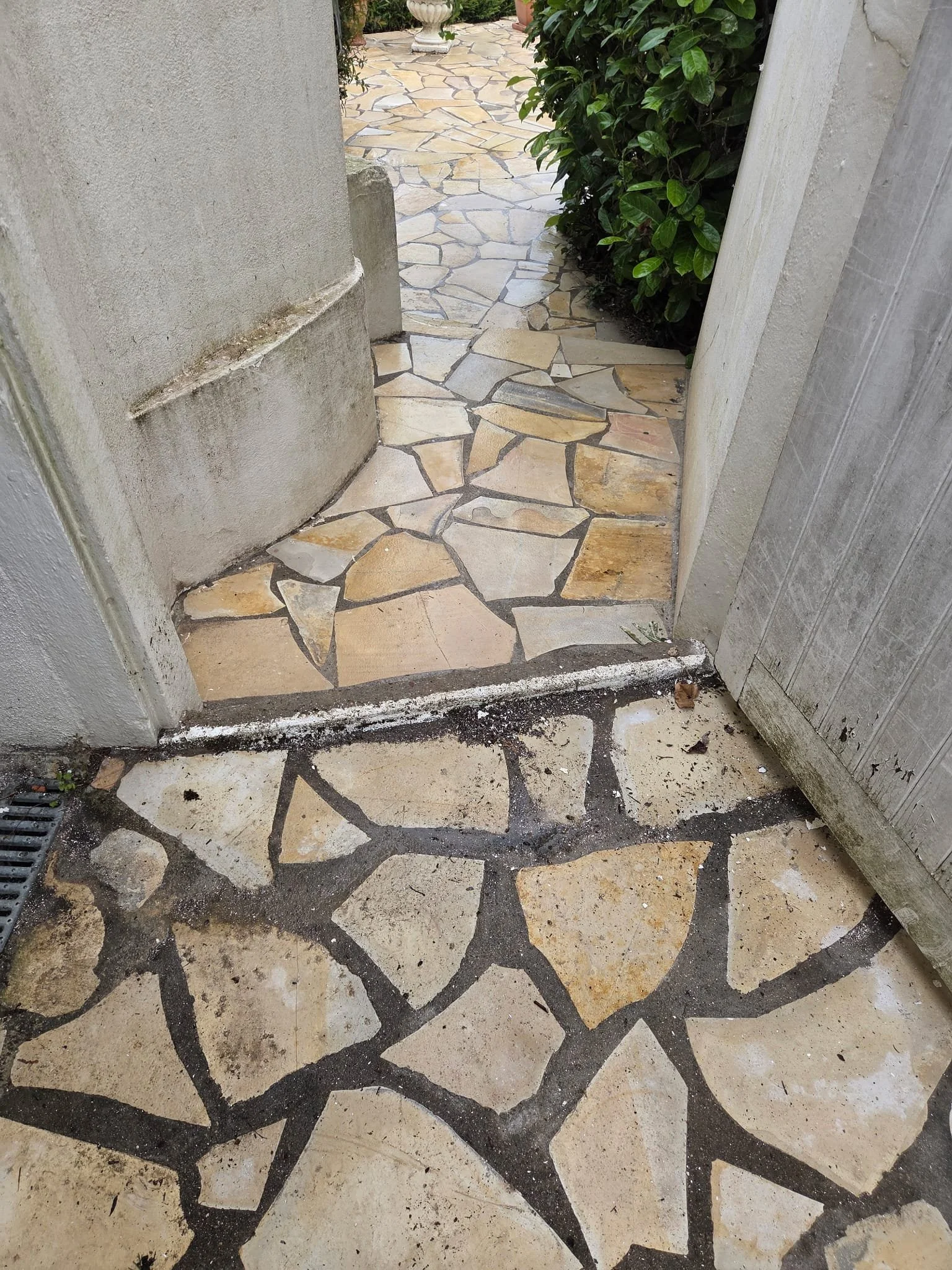 Un passage en pierre avec un sol en carreaux de pierre beige et un mur en bois grisâtre, entouré de verdure.