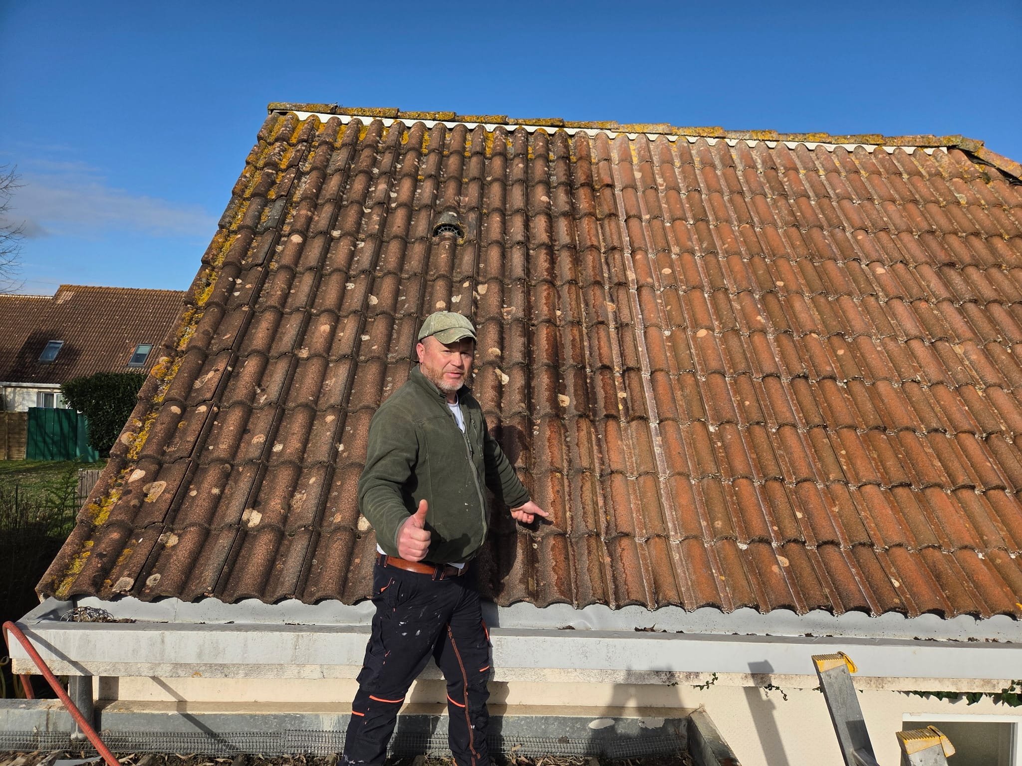 Un homme avec une casquette donne un pouce en l'air devant un toit en tuiles rougeâtres, en cours de rénovation ou d’entretien.