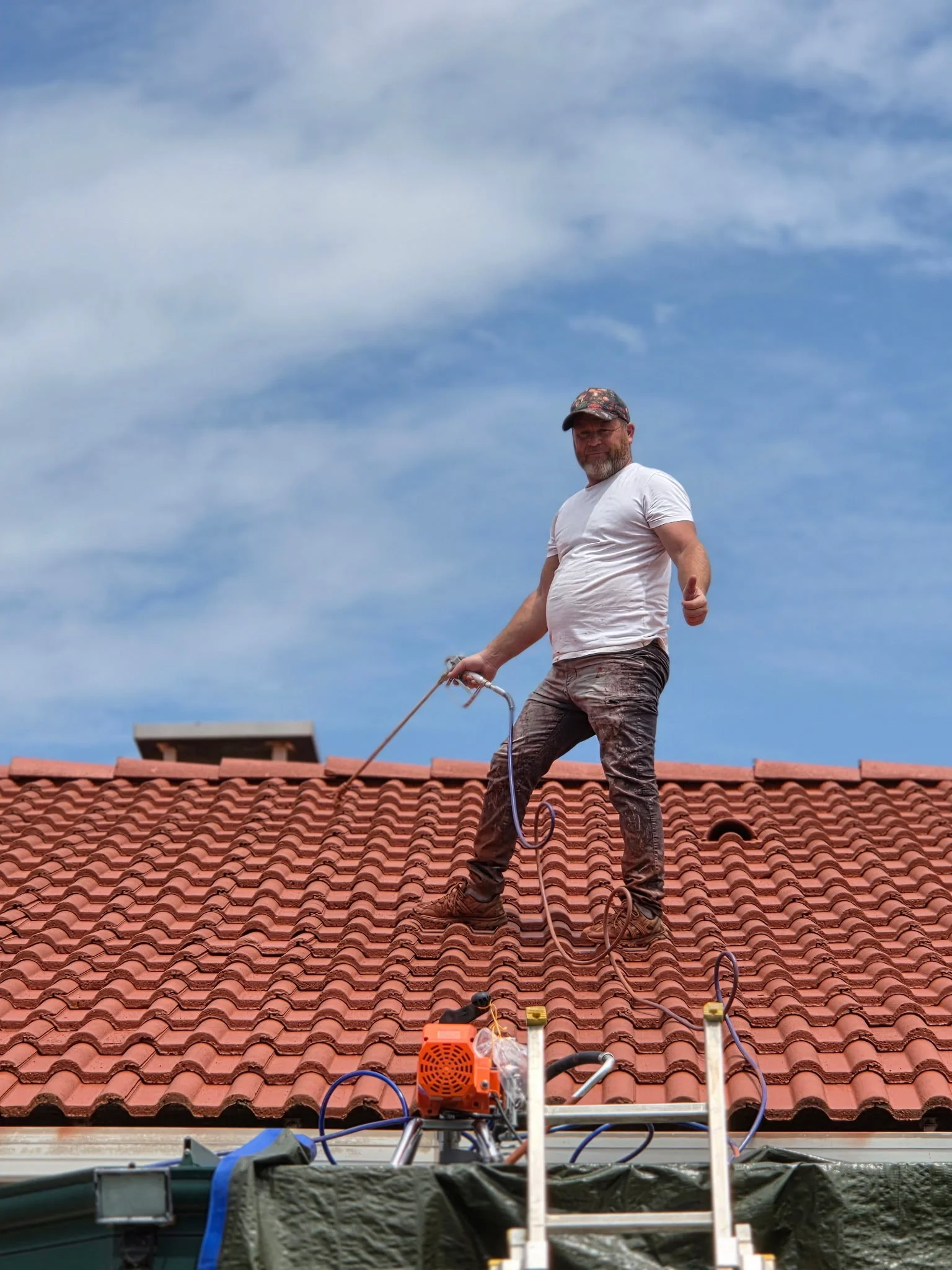 Un homme travaille sur le toit d'une maison avec des tuiles rouges, utilisant des équipements de sécurité et un outil électrique, sous un ciel bleu avec quelques nuages.