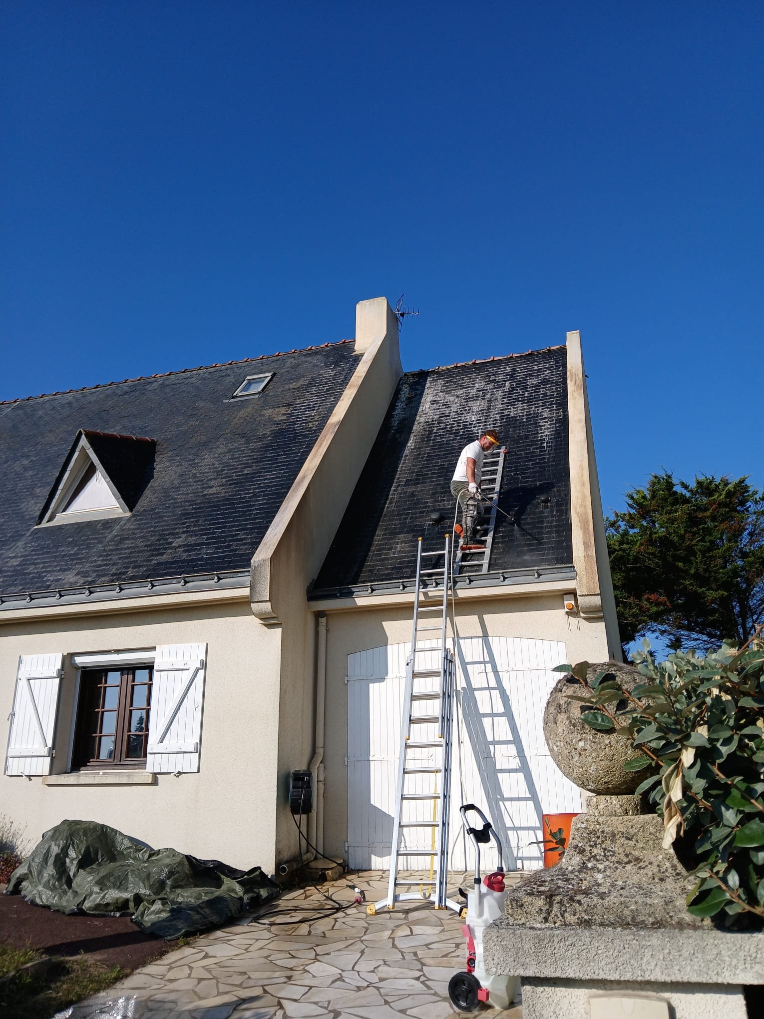 Un homme en train de nettoyer ou de réparer le toit d'une maison à l'aide d'une échelle, sous un ciel bleu clair.