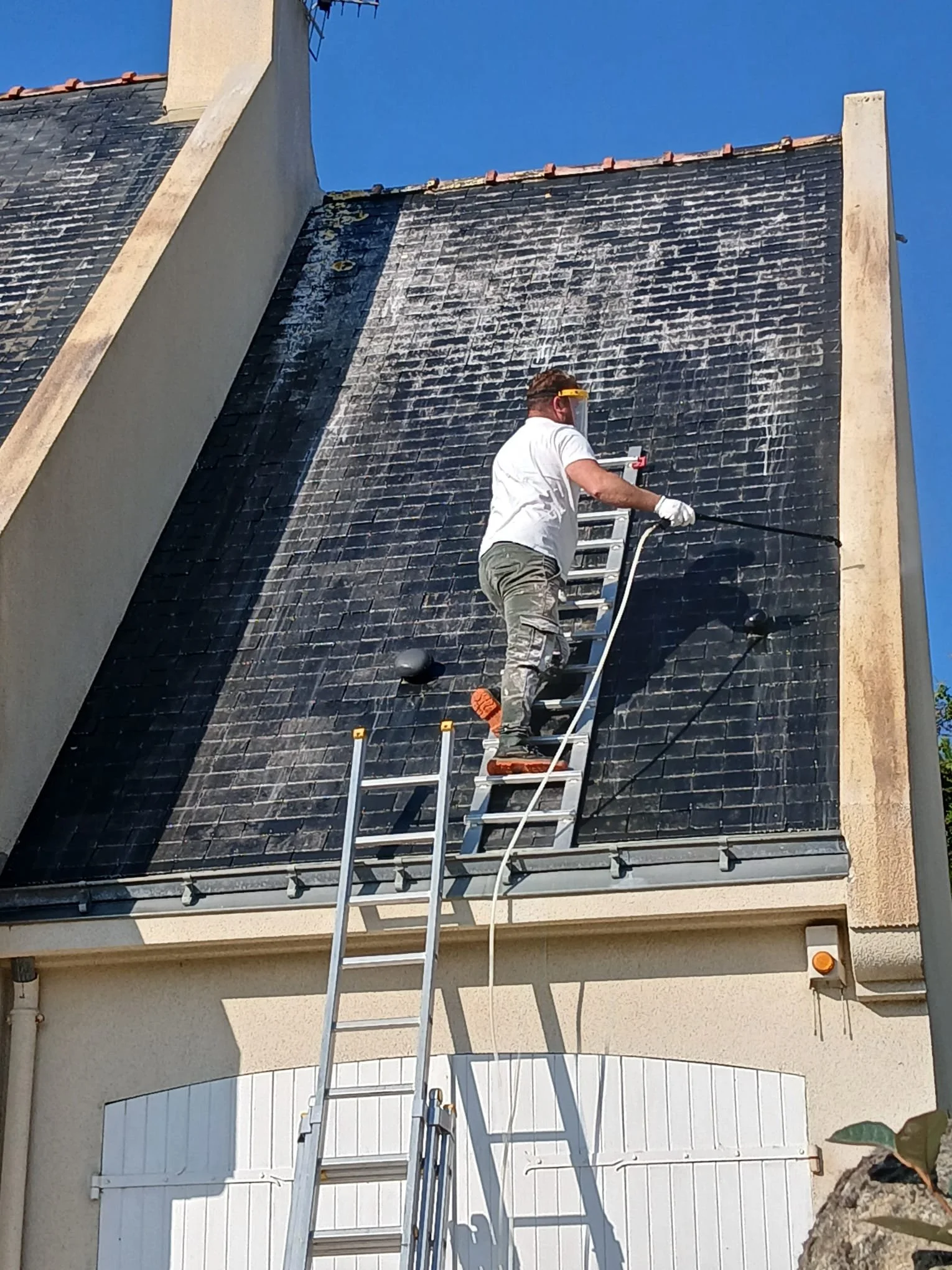 Un homme en haut d'une échelle nettoie la toiture en ardoise d'une maison avec un jet d'eau. Il porte des gants, un t-shirt blanc et un casque de protection.