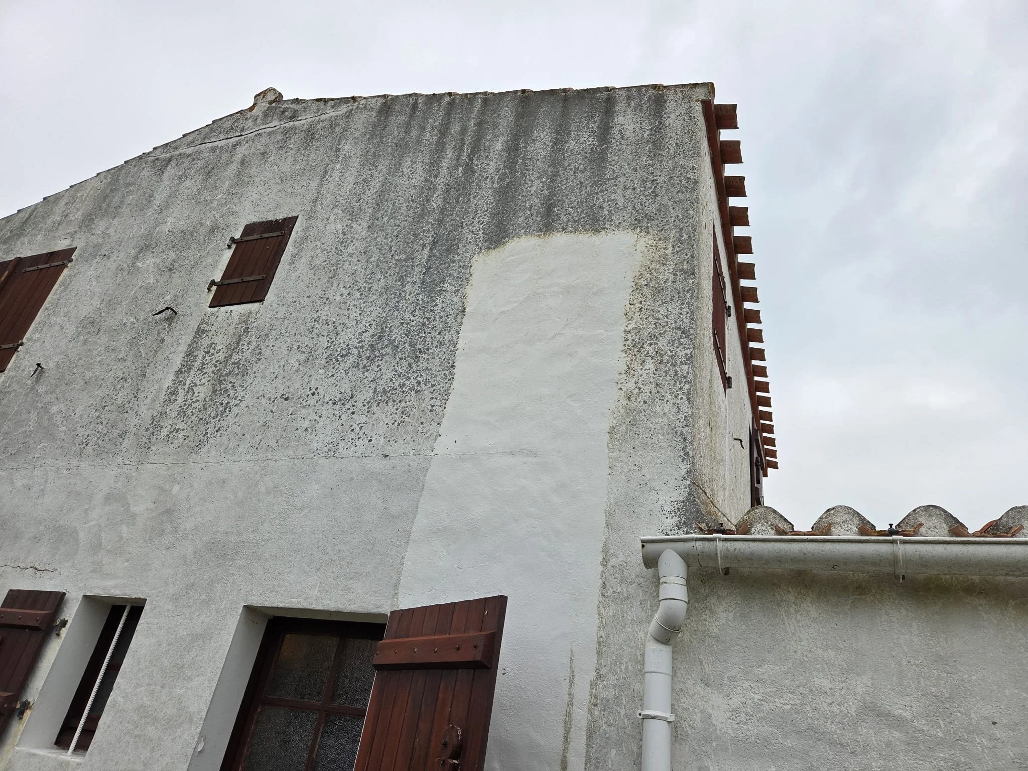 Vue d'un bâtiment résidentiel blanc avec fenêtres à volets en bois fermés, tuyaux de plomberie visibles, et un toit en tuiles. Le ciel est gris.