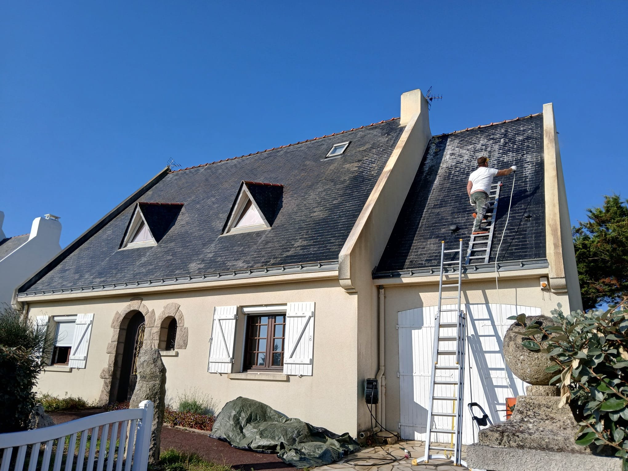 Un homme en t-shirt blanc nettoie le toit en ardoise d'une maison à l'aide d'une échelle. La maison a des fenêtres avec volets blancs et un mur en pierre au niveau de l'entrée. Le ciel est clair et bleu.