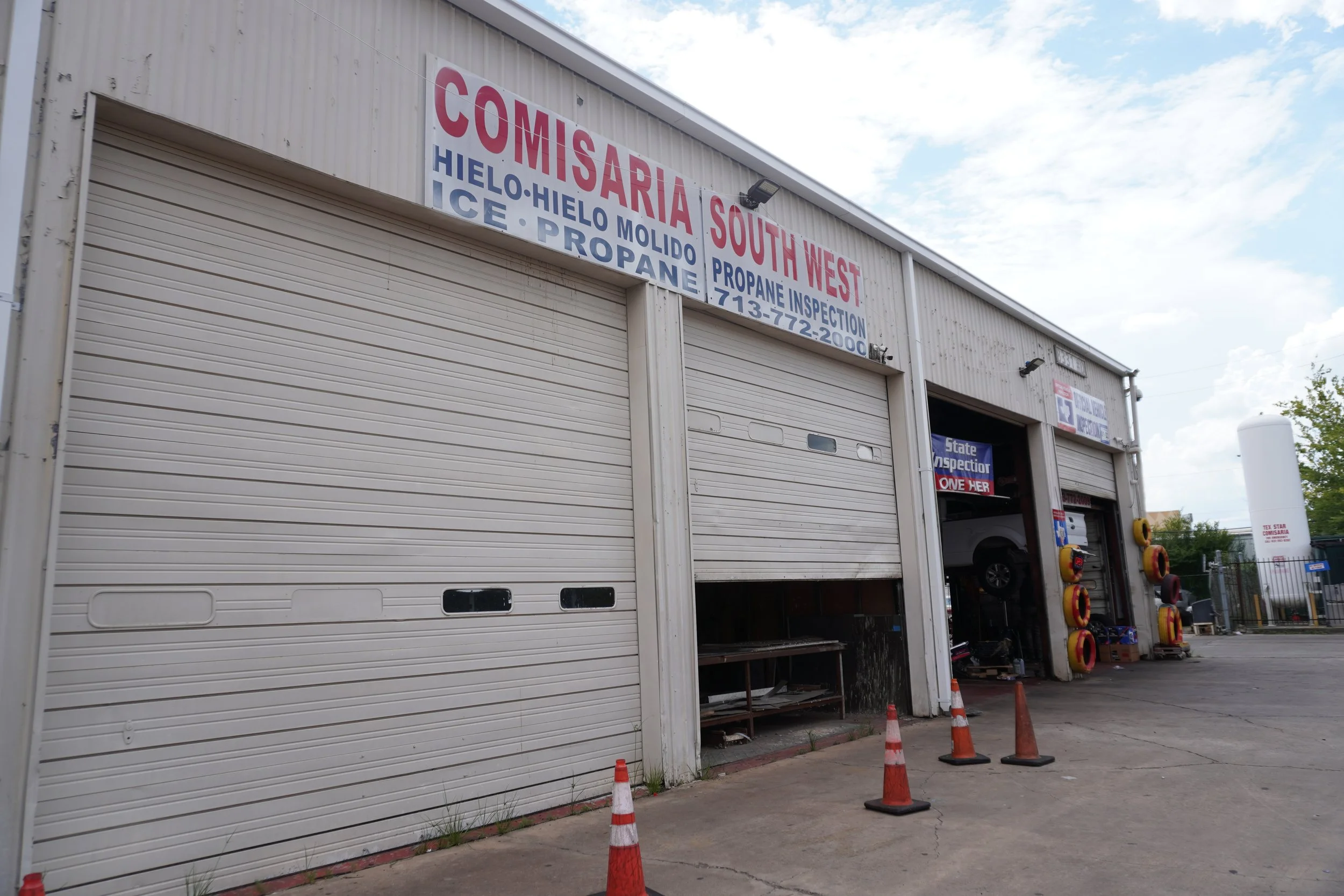 Exterior of a building with a sign for a wholesale ice and propane business named 'Comisaría South West.' The sign also mentions ice, propane, and inspection services with a phone number. The building has two garage doors, with some open sections showing equipment and supplies inside. Orange traffic cones are placed in front of the building.