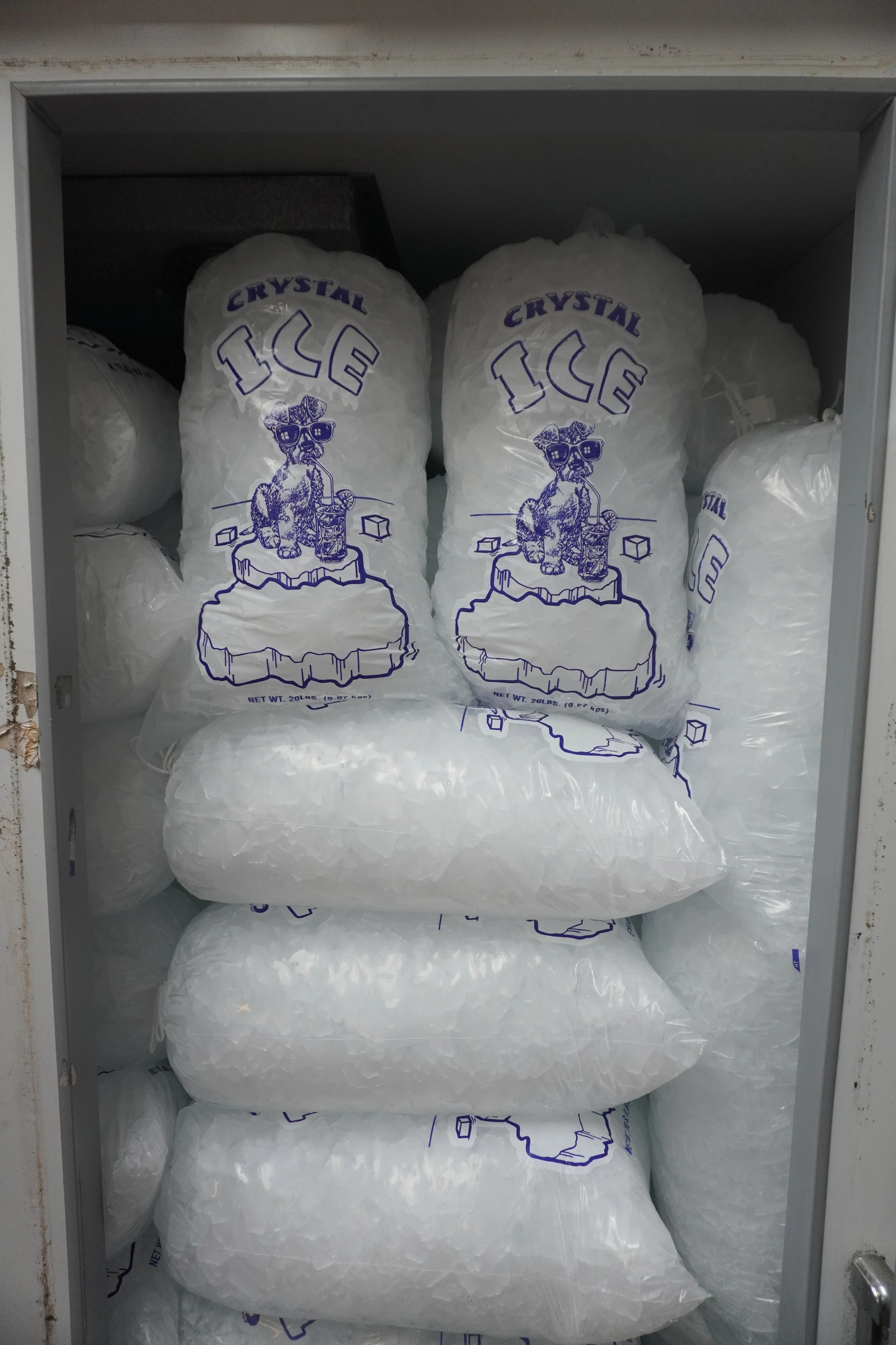 Bathroom storage cabinet filled with multiple white bags of ice labeled "CRYSTAL ICE" with a cartoon dog wearing sunglasses on the bags.