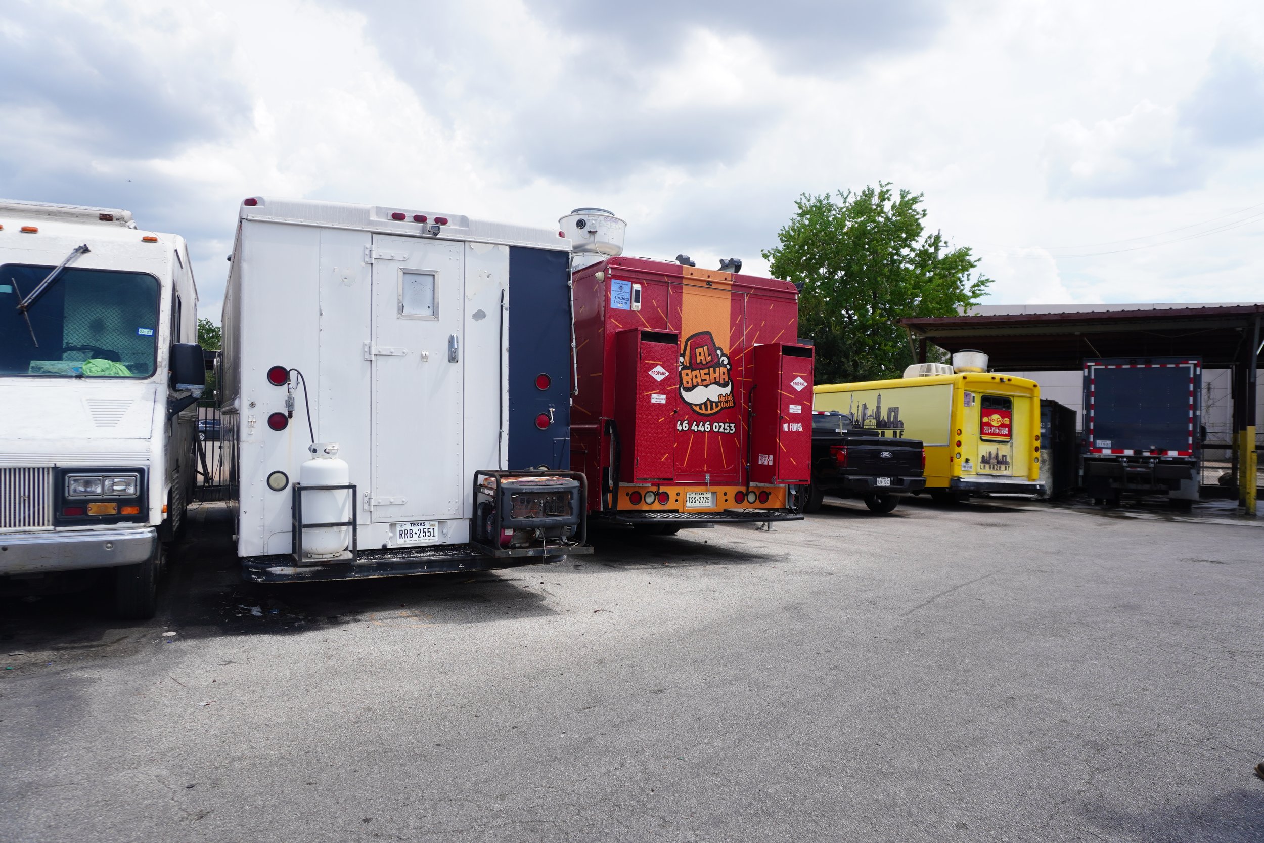 A row of food trucks and parked vehicles at an outdoor location under a cloudy sky, including a white food truck, a red and orange food truck with a logo, a yellow truck, and a black truck, with a yellow post and a shed in the background.