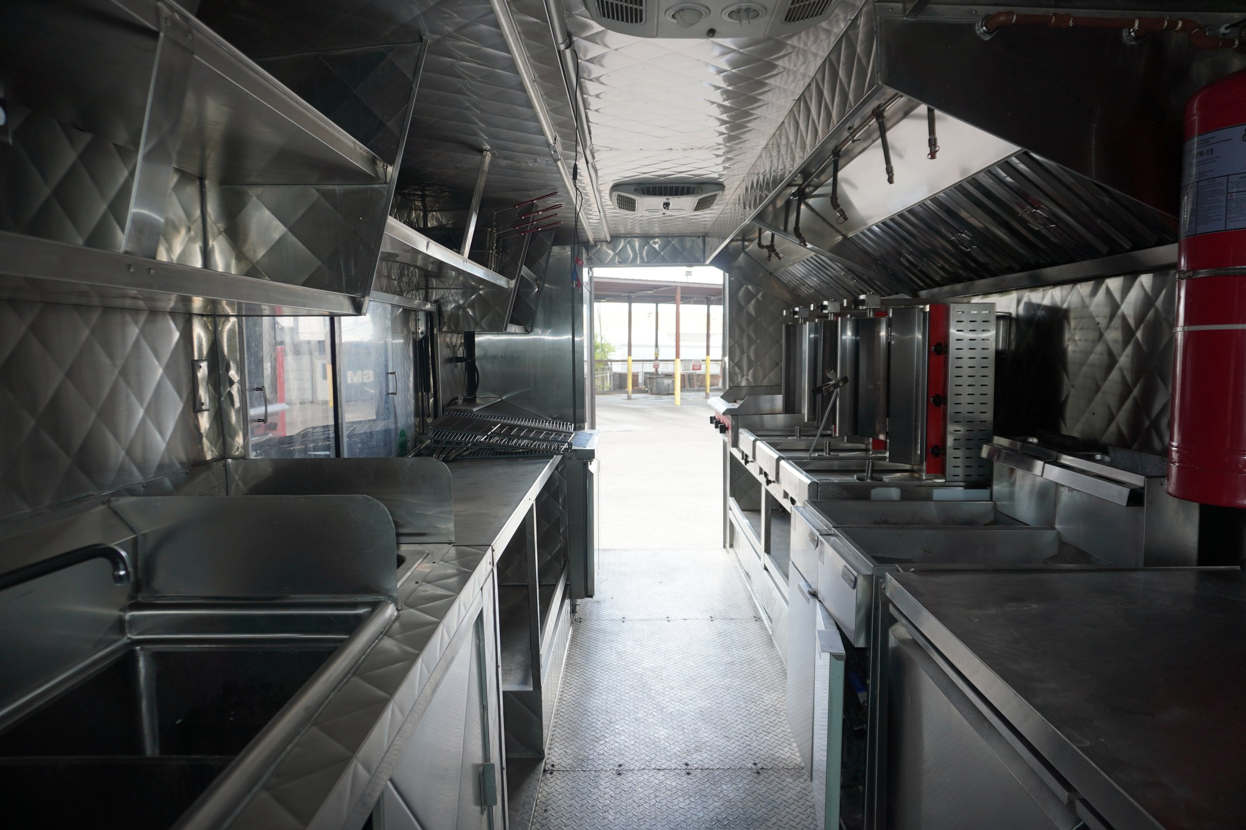 The interior of a food truck kitchen with stainless steel countertops, appliances, and storage shelves. There is a window at the back opening to the outside, showing some yellow bollards and a parking lot.