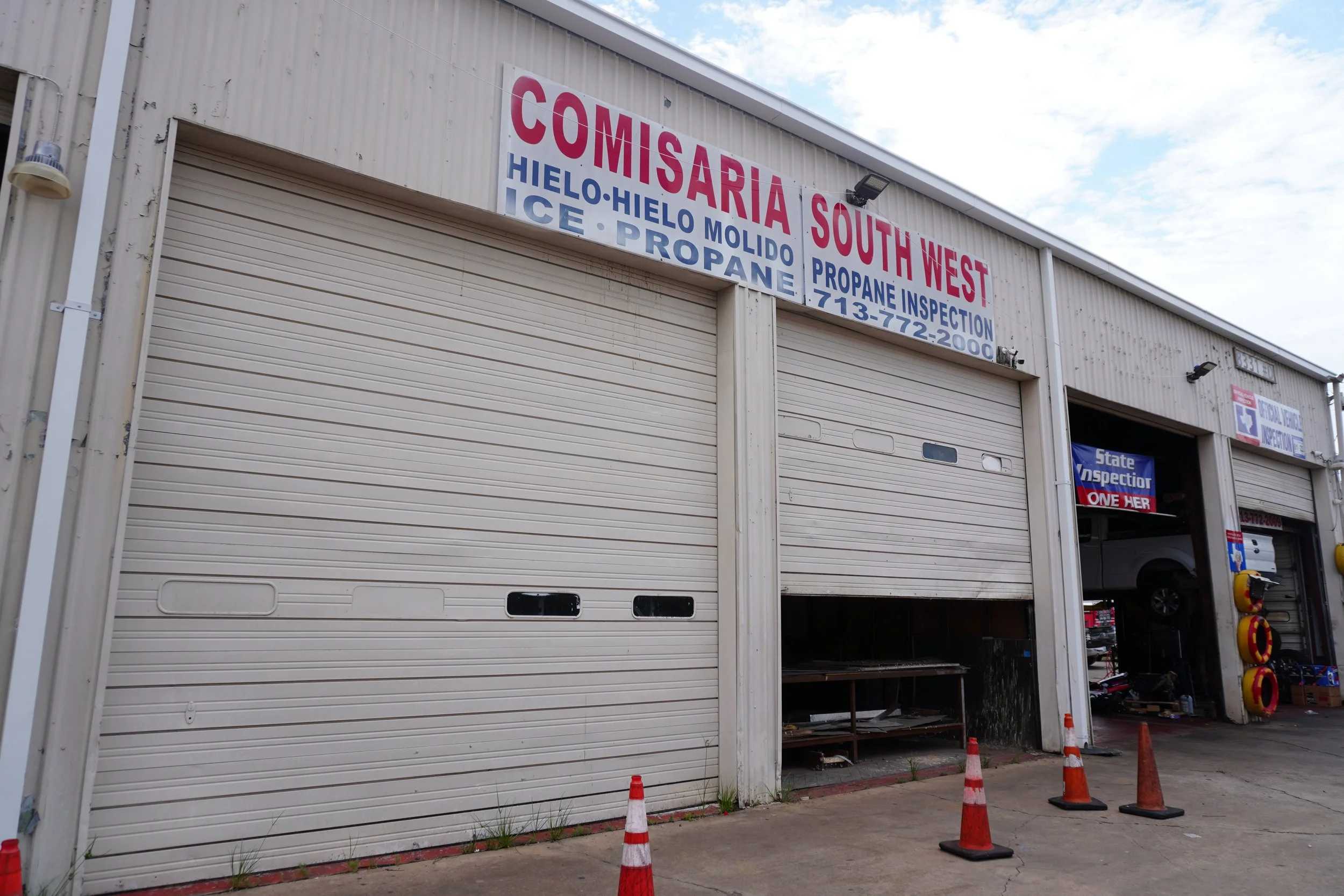 Exterior of a building with a sign indicating it is a propane and ice supply store, with several orange traffic cones outside and part of a garage door open.