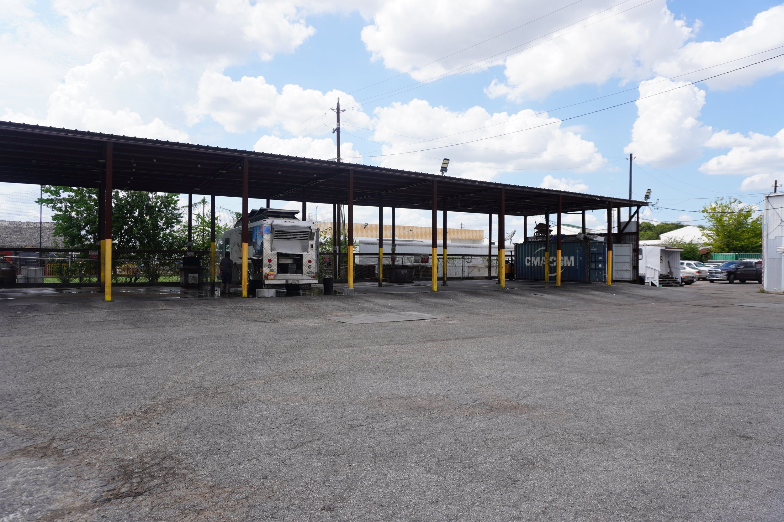 Empty parking lot with a covered food vending area, a food truck, and a blue shipping container, under a partly cloudy sky.