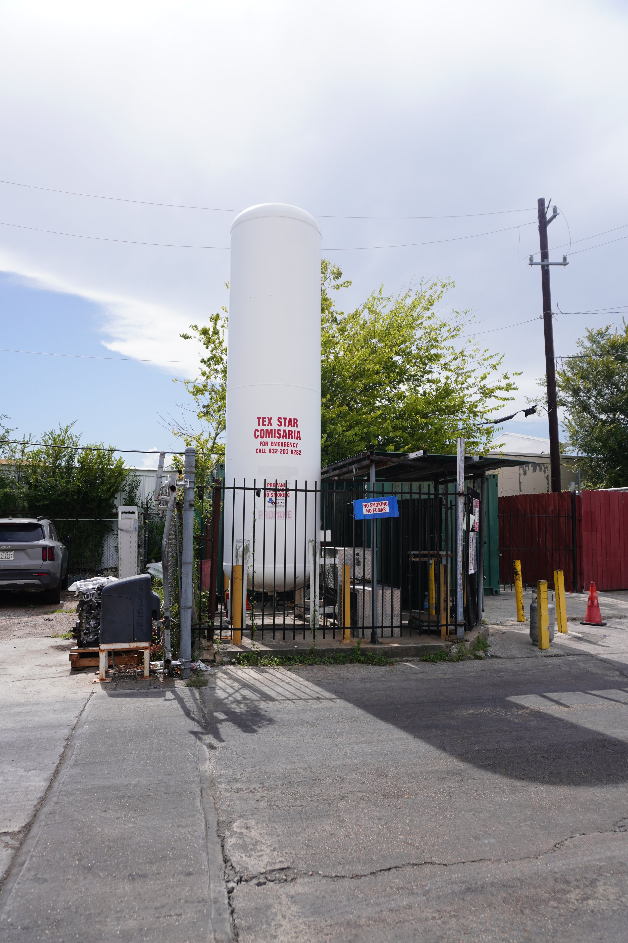 A utility station with a tall white propane tank labeled 'TEX STAR COMISARIA.' The station is enclosed by a metal fence and has signs that say 'No Smoking' and 'Prohibited' in Spanish. There are trees, power lines, and parked cars around the station, with a partly cloudy sky overhead.