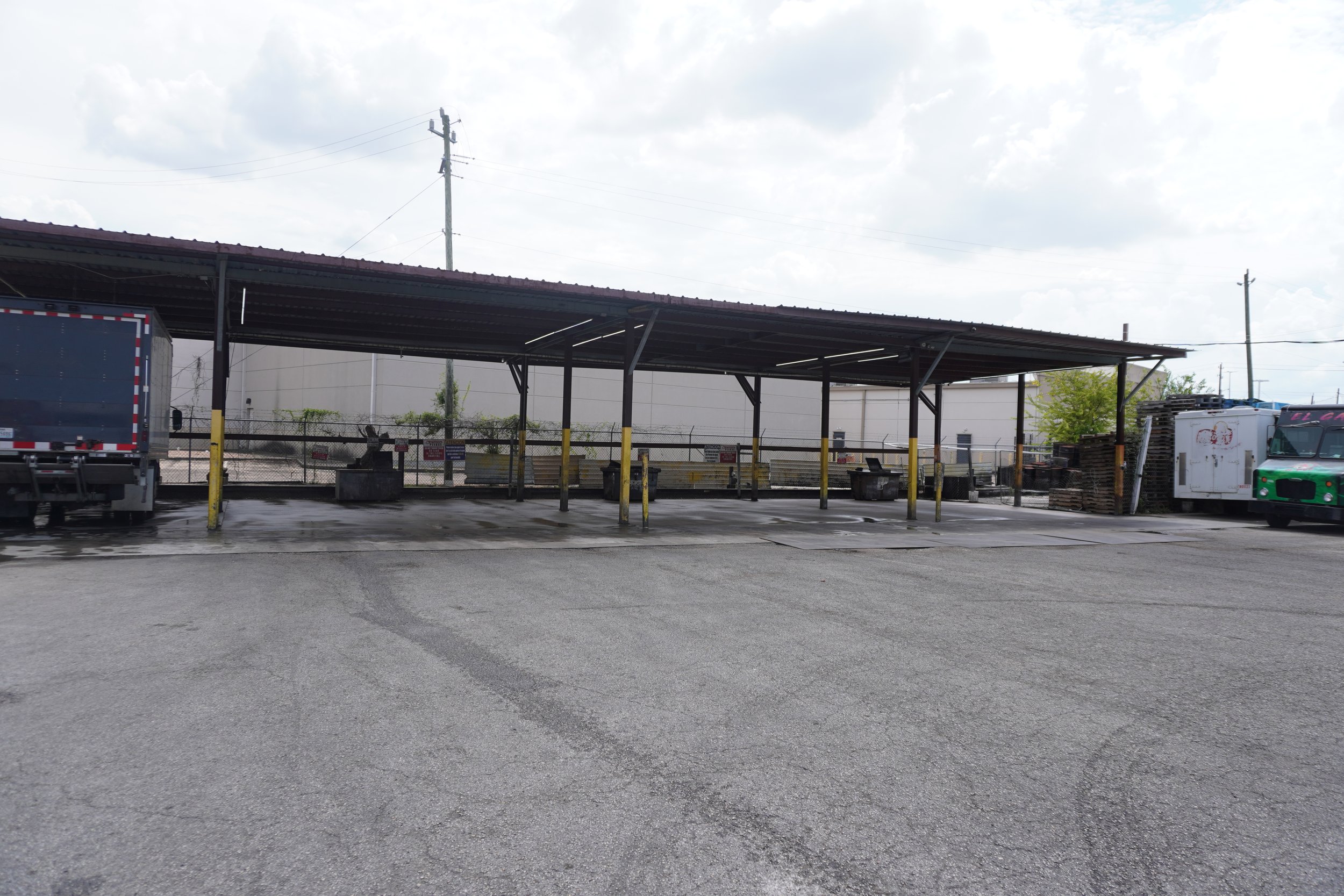 Empty parking lot with a covered structure, some trucks, and storage pallets, under a cloudy sky.