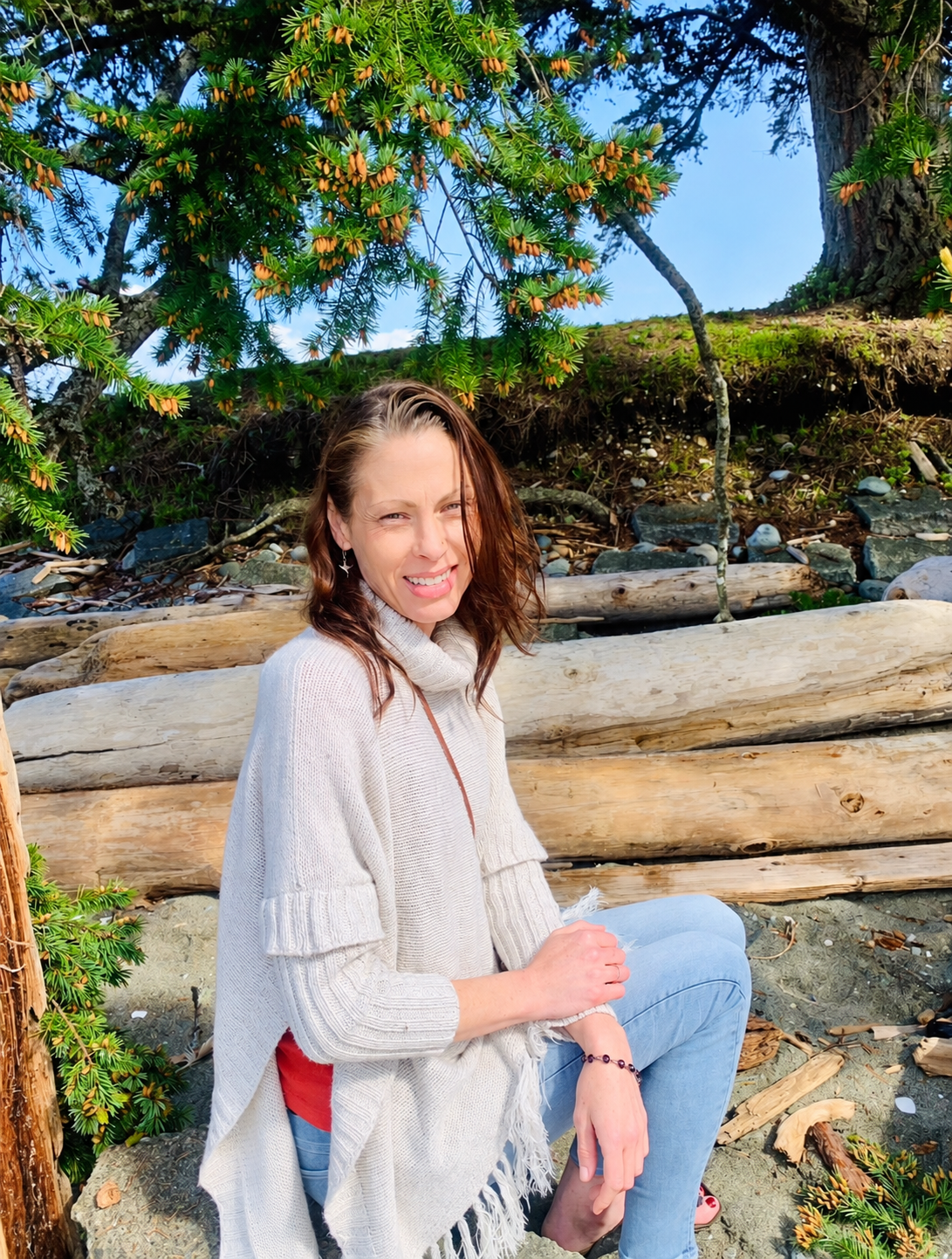 Joanna Lumley seated on driftwood beneath evergreen trees.