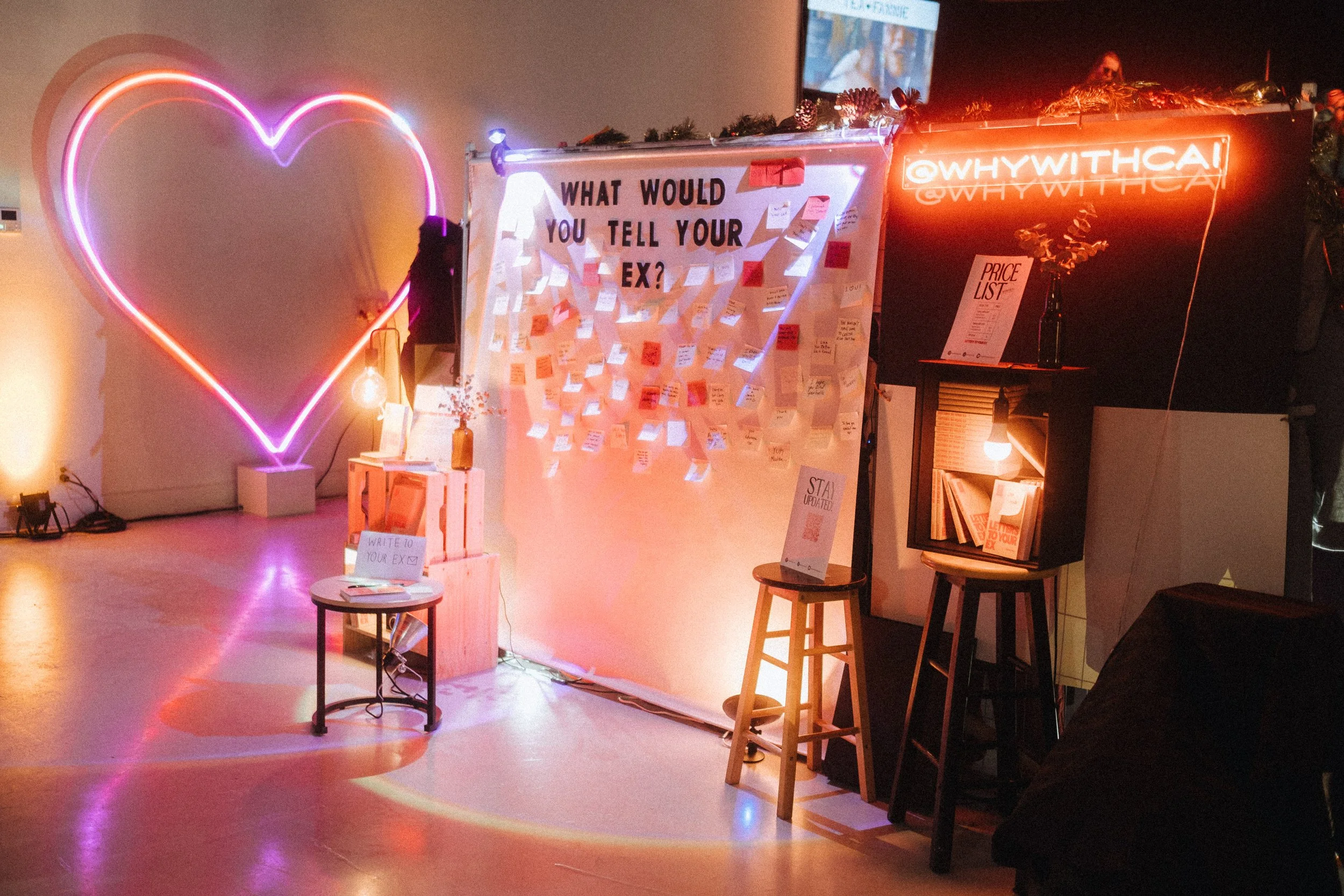 A cozy, dimly lit space with neon lights and a whiteboard asking 'What would you tell your ex?' covered with sticky notes, a neon pink and purple heart on the wall, a small table with a sign saying 'Write to your ex,' and various seating and decorative elements.